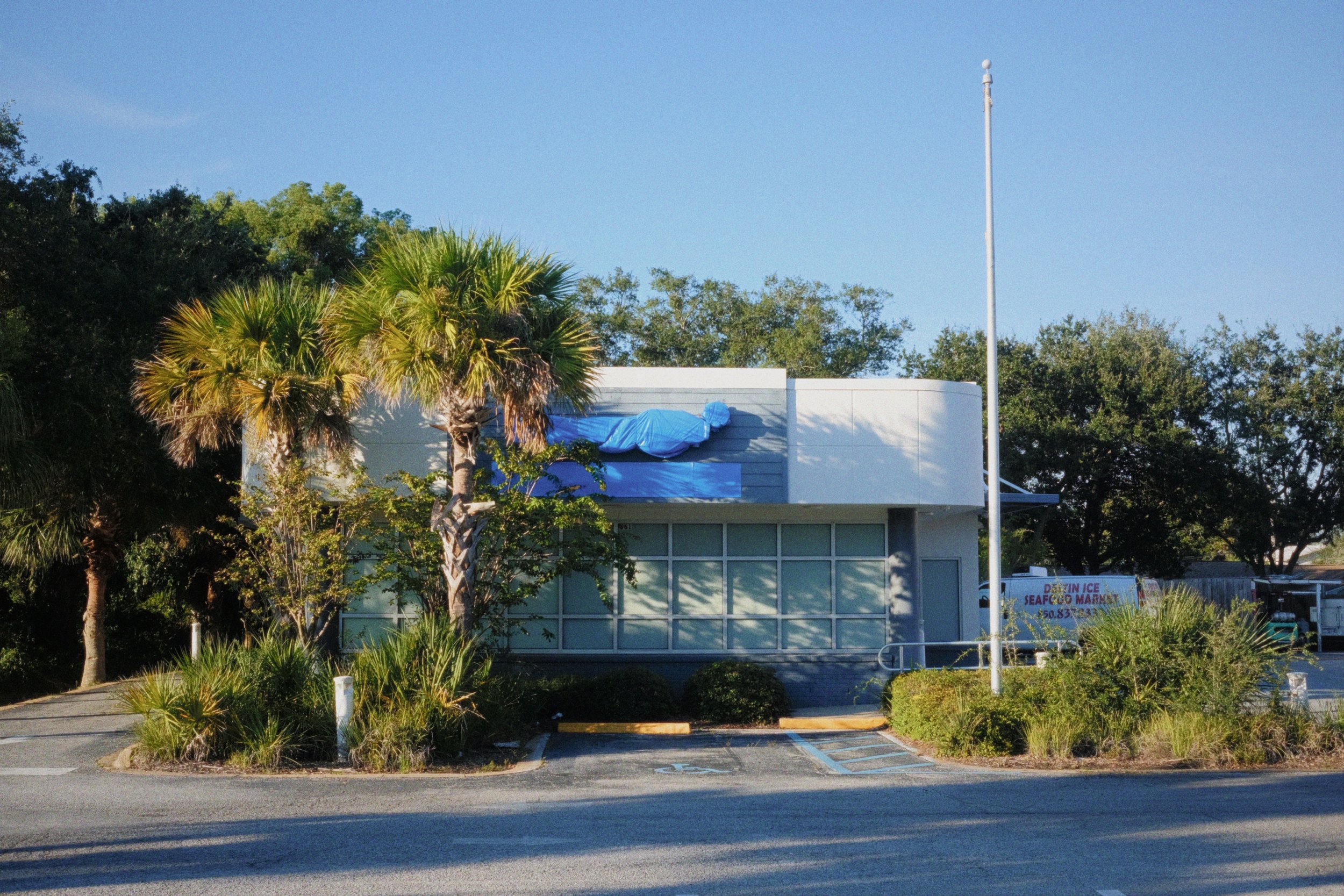 Small building with large windows, surrounded by trees and plants, with a parking lot in front and a flagpole to the right. Blue sky overhead.