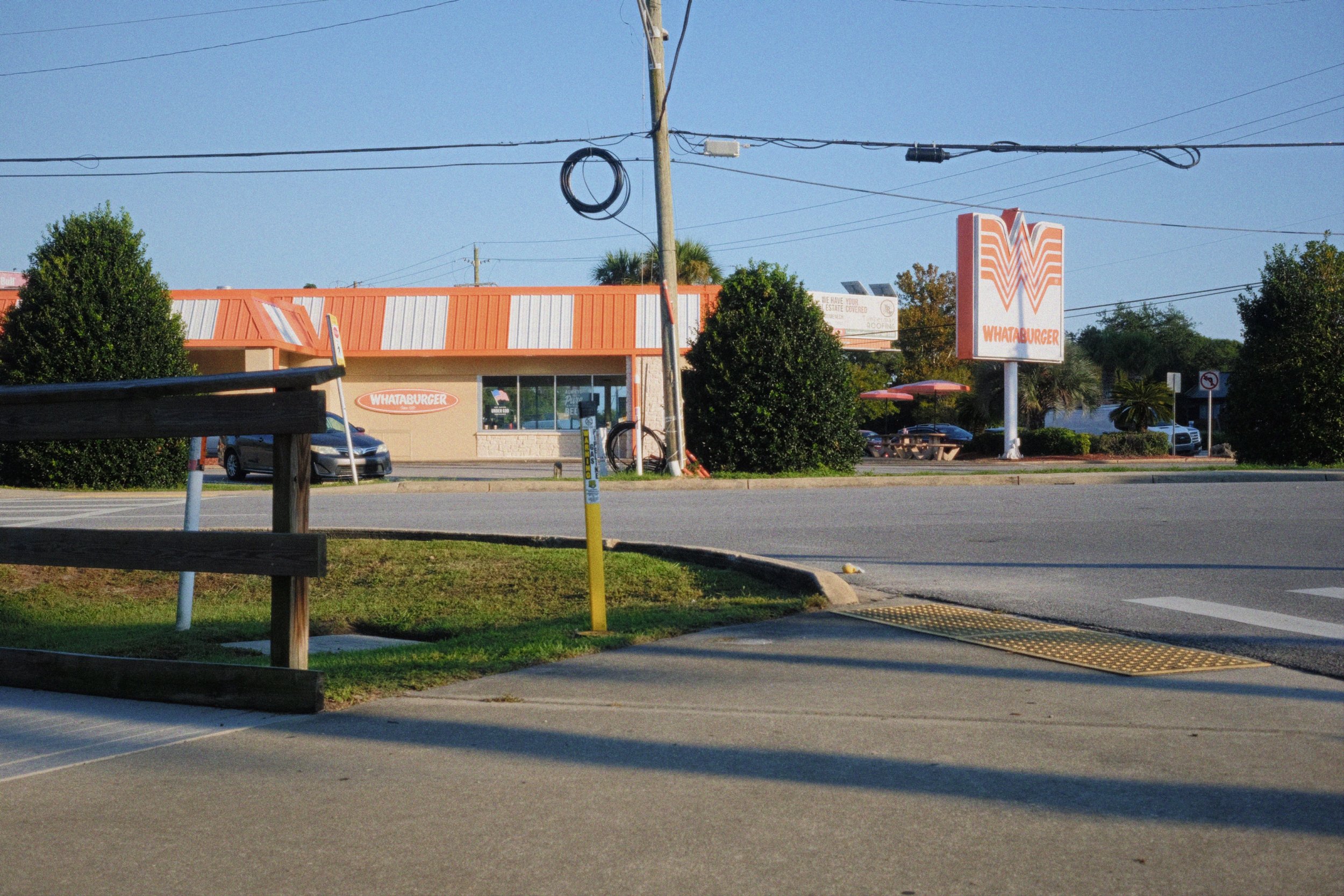 A parking lot in front of a Whataburger restaurant with trees, utility poles, and power lines under a clear blue sky.