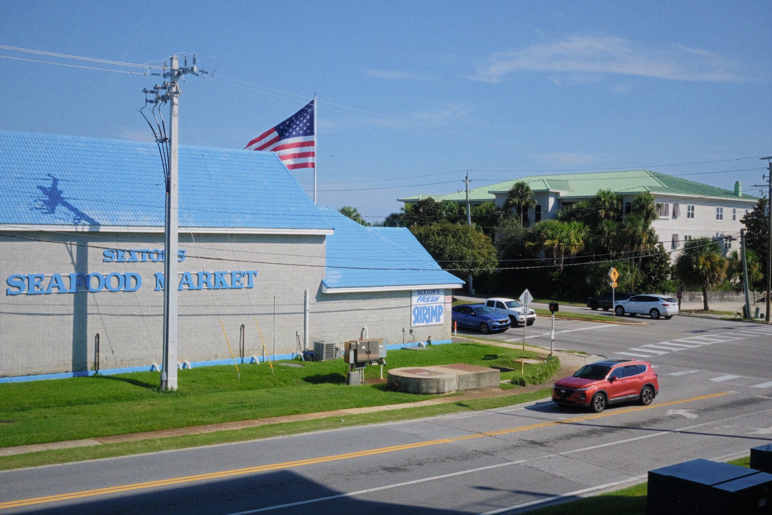The image shows a seafood market building with a blue roof. An American flag is flying on a pole. There are palm trees and other buildings in the background, along with several cars.