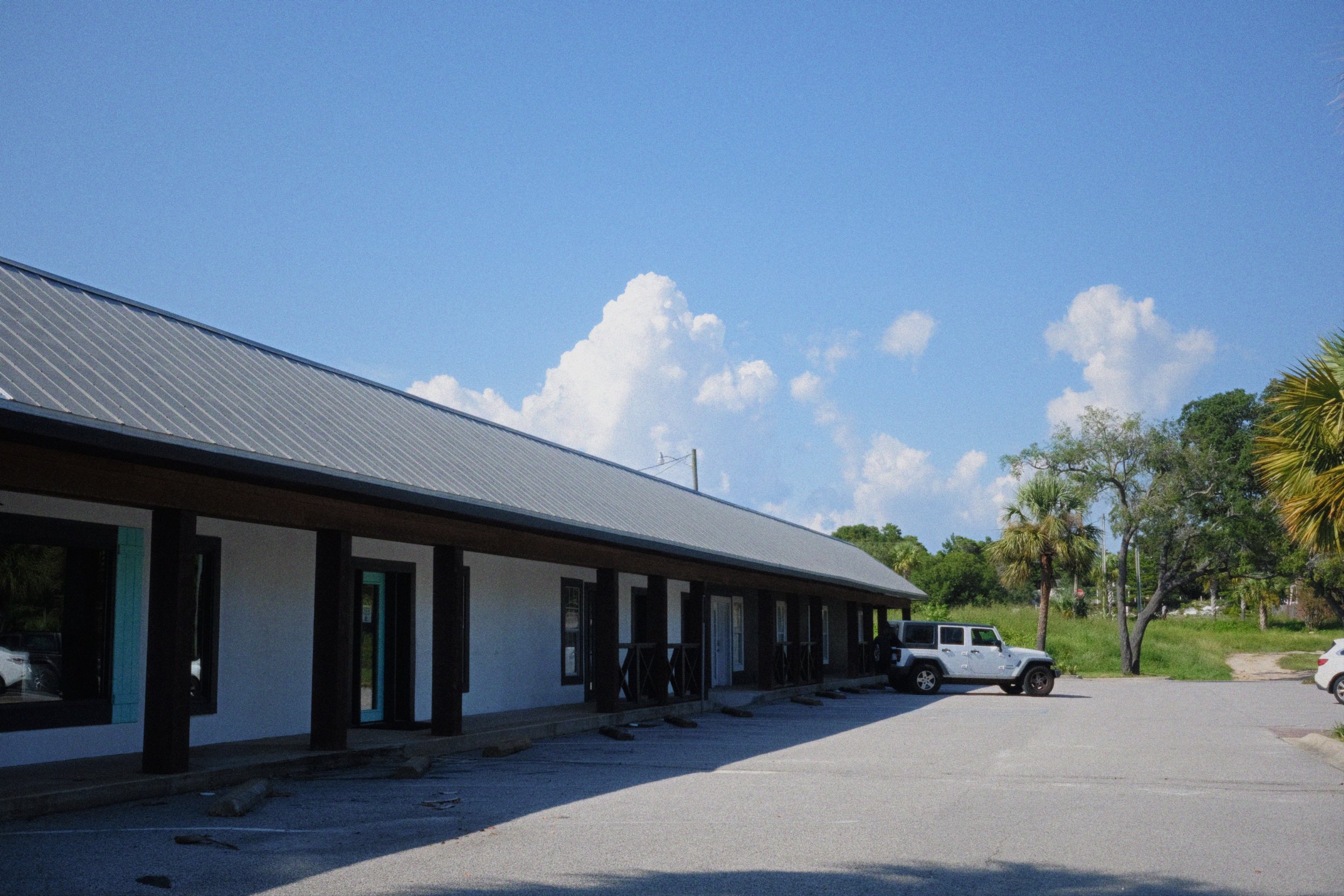 A building with a metal roof, parked cars, trees, and a partly cloudy blue sky.