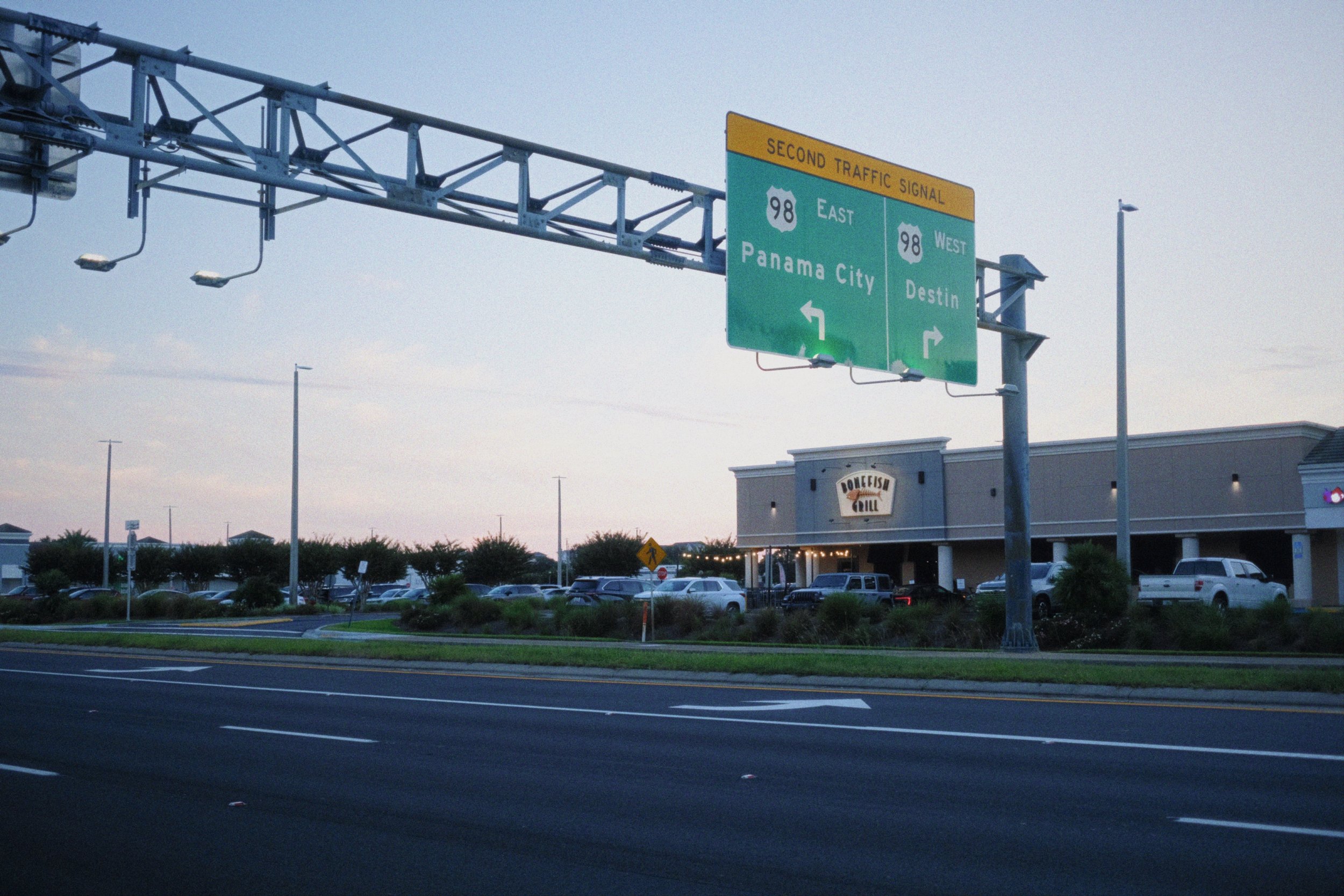 Highway road sign indicating directions to Panama City East US Highway 98 east and Destin via US Highway 98 west, with a shopping plaza and parking lot visible in the background at dusk.