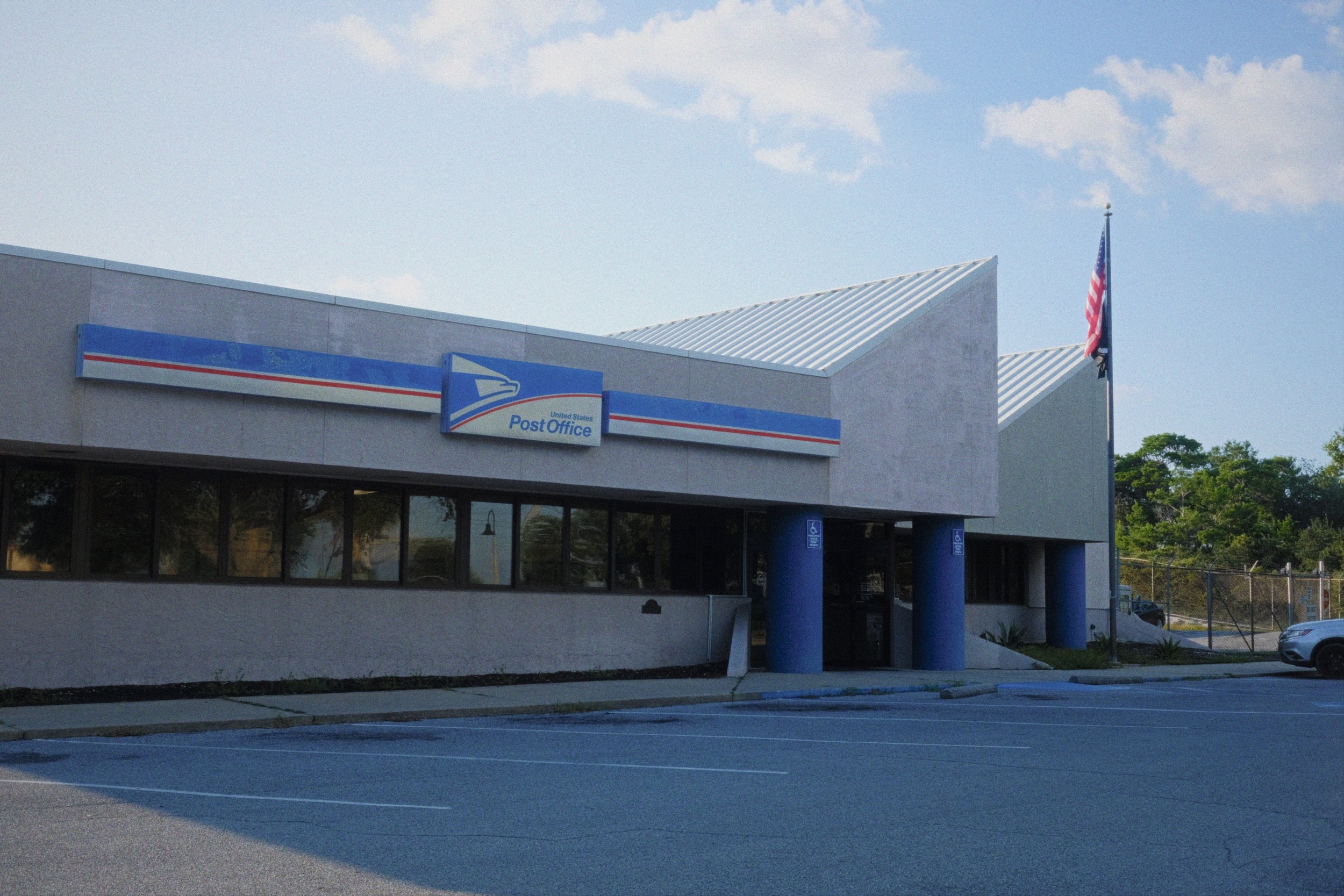 United States Post Office building with a sign, an American flag, and parking lot in front.