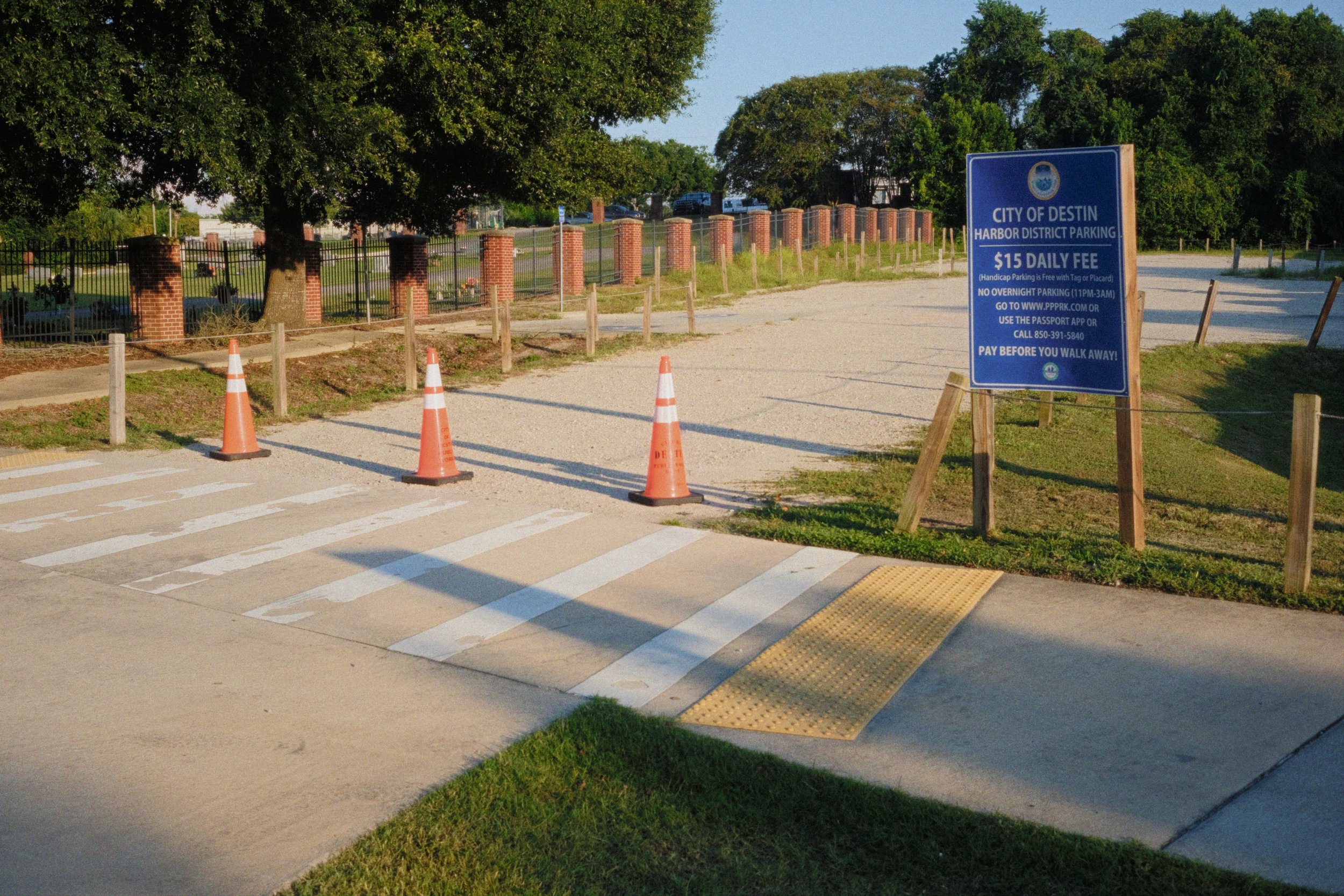 Parking lot with orange safety cones and a blue informational sign about parking fees, surrounded by trees and a brick fence.
