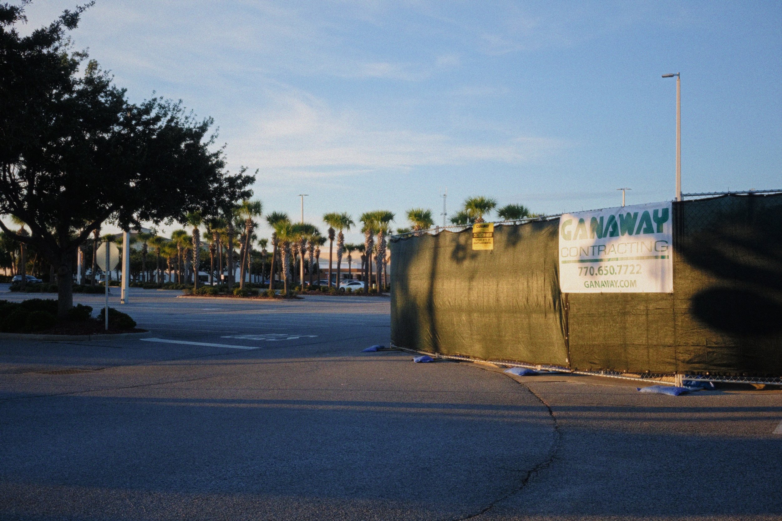 Empty parking lot with trees and a construction barrier displaying an outdoor advertisement for Ganaway Contracting, with a partly cloudy sky in the background.