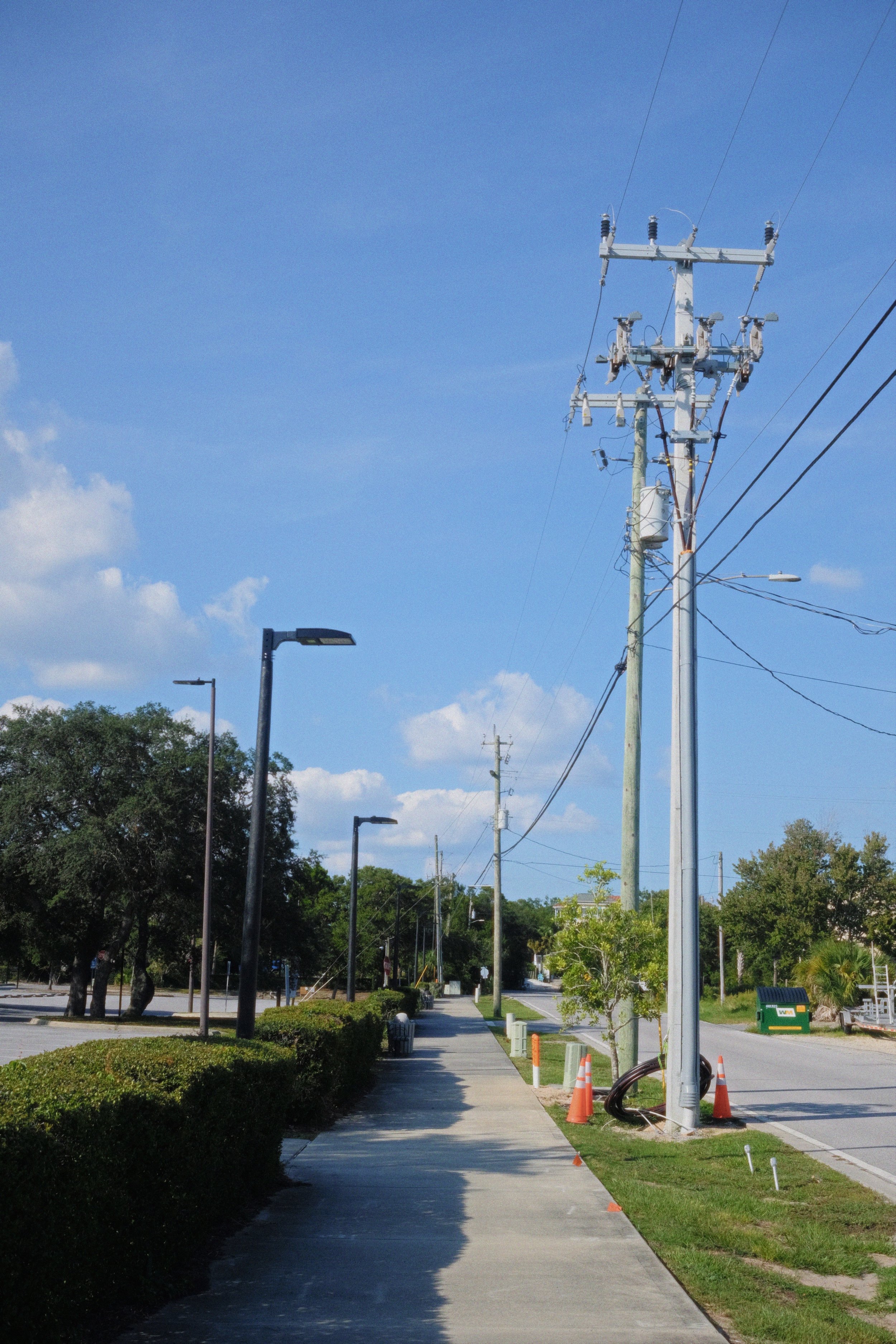 A sidewalk along a parking lot with street lamps and trees, close to a utility pole with overhead wires, some orange traffic cones and a green trash bin, under a blue sky with light clouds.