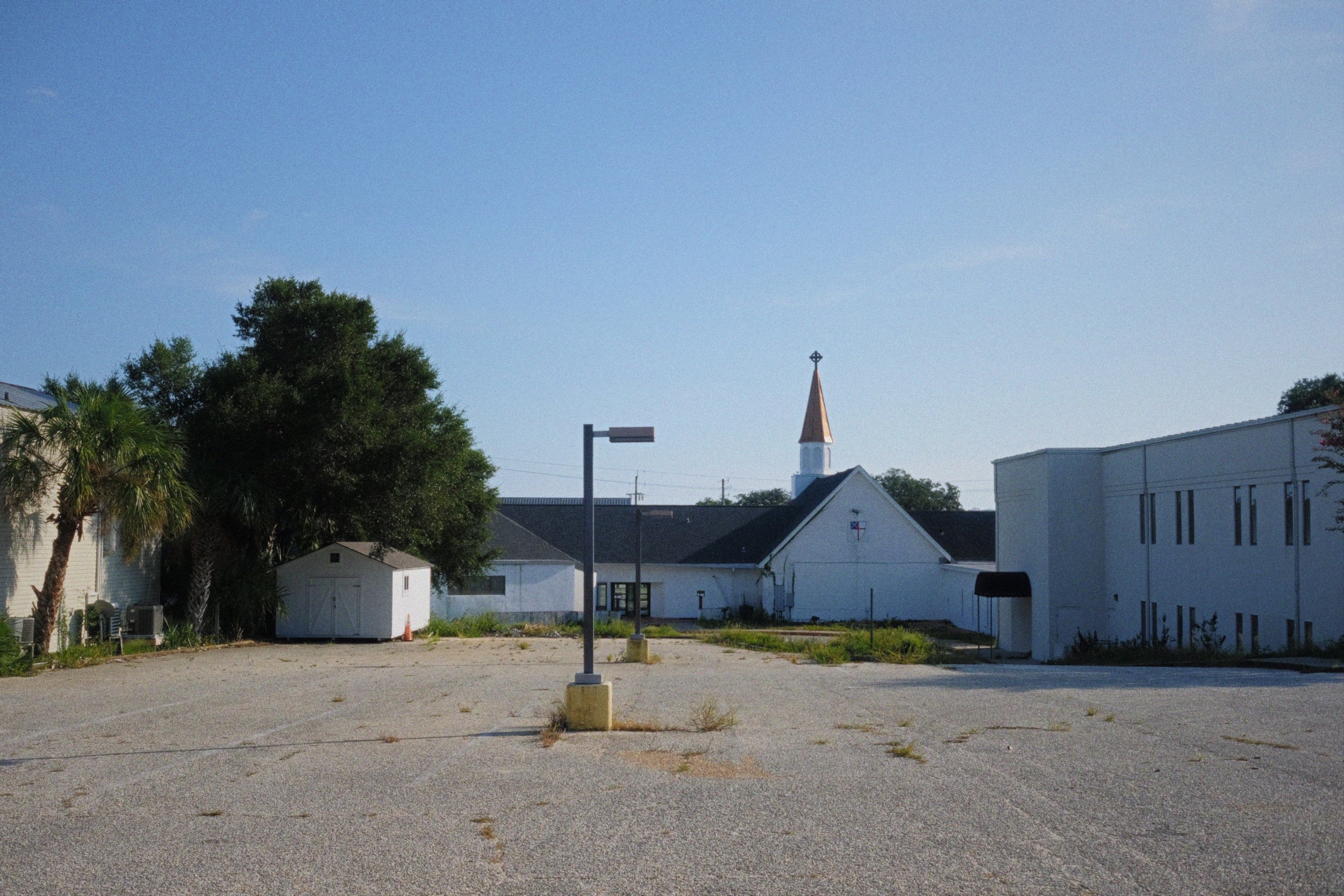 Empty parking lot with a small white building, a church with a steeple, trees, and a clear blue sky in the background.
