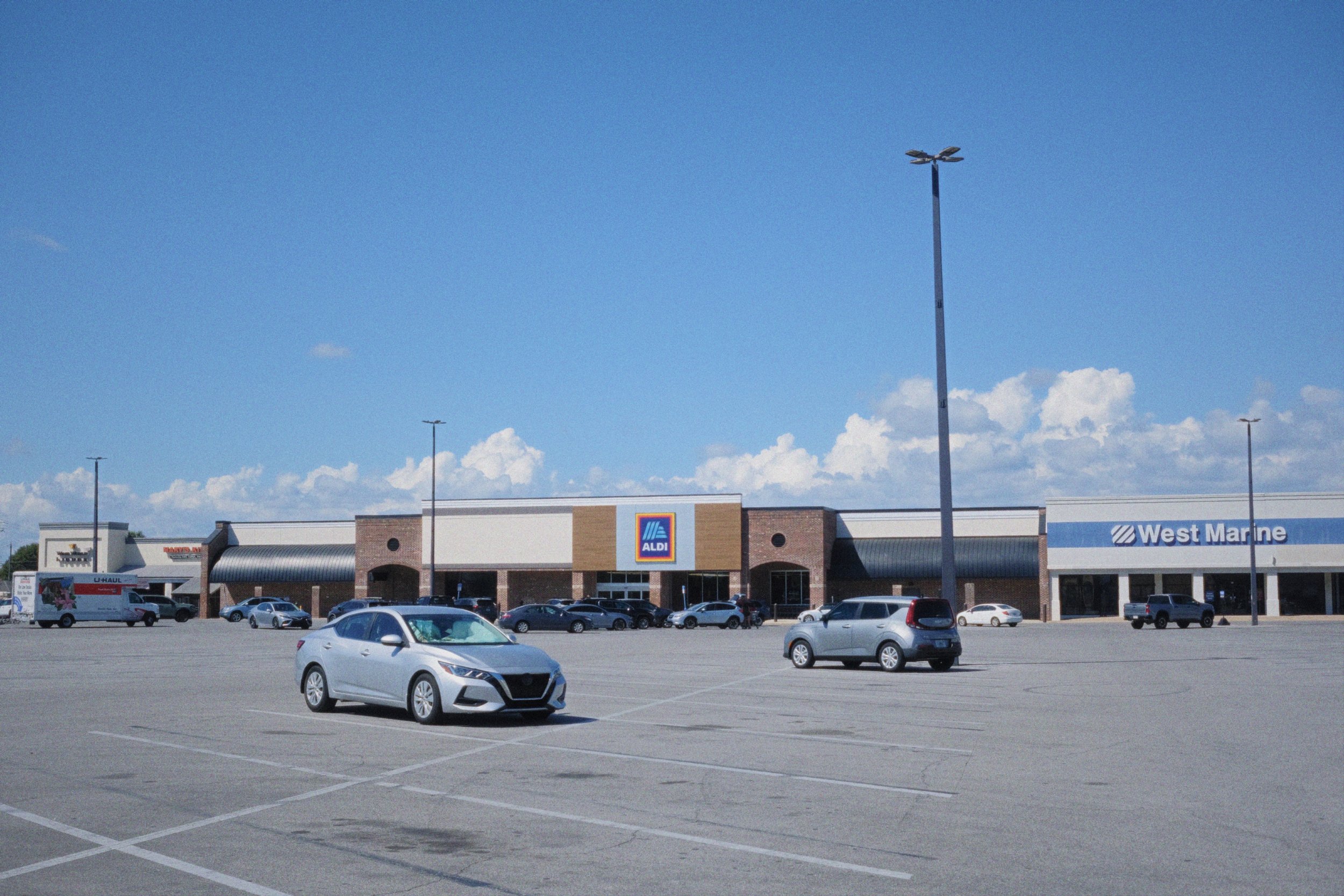 A parking lot with several cars in front of a shopping center. The shopping center has stores including ALDI and West Marine. The sky is blue with some clouds.