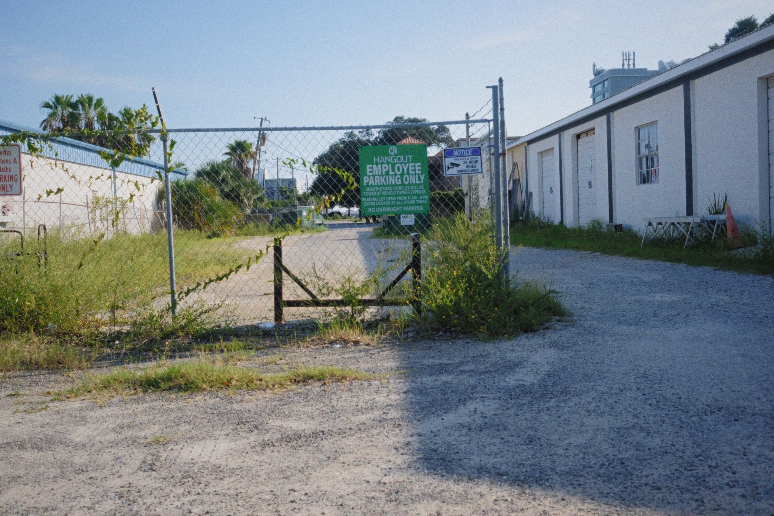 View of an outdoor parking area with a chain-link fence, signs indicating employee parking and video surveillance, and buildings and palm trees in the background.