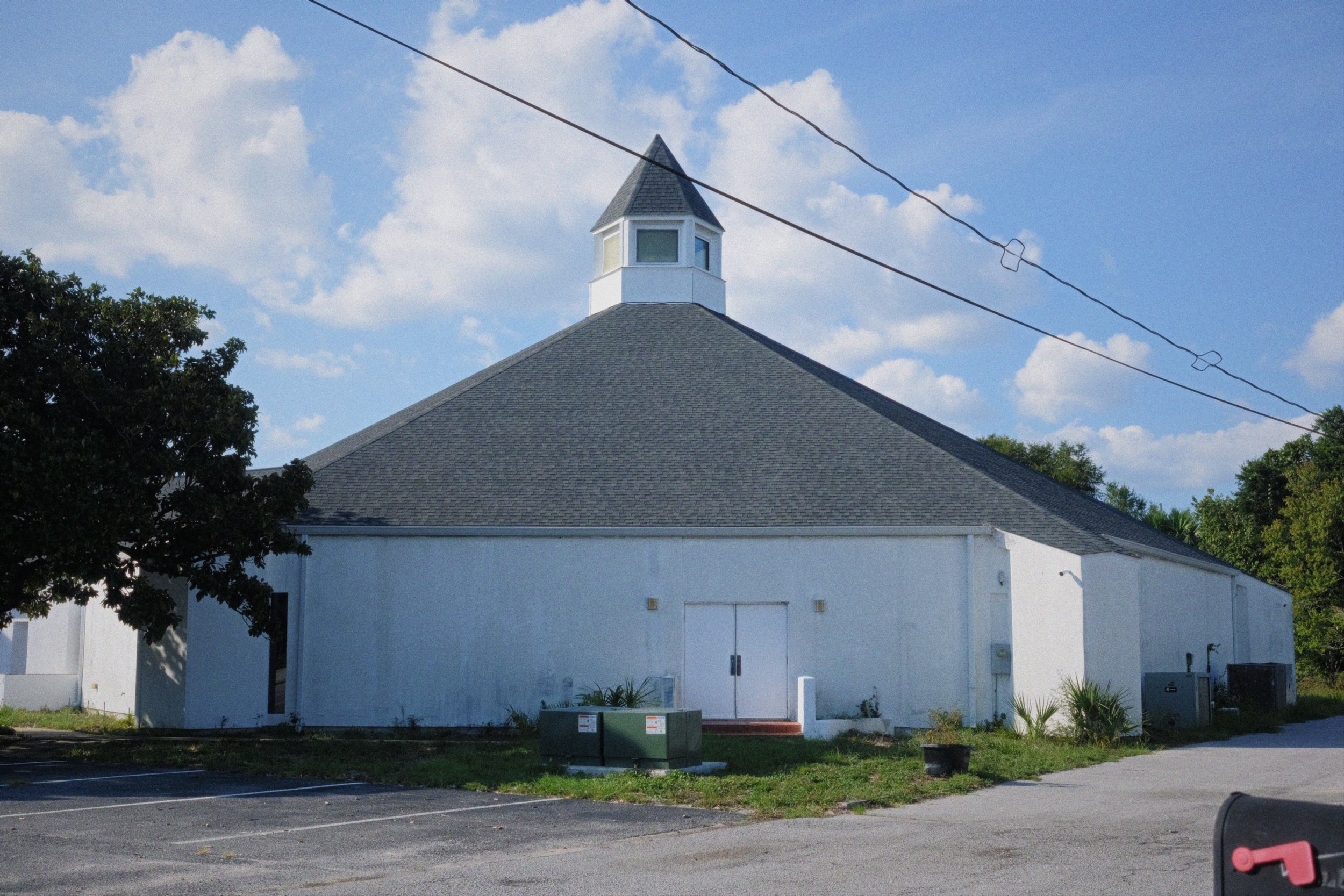 A white building with a pointed roof and a small tower with windows on top, set against a blue sky with scattered clouds. Trees surround the building, and power lines are visible overhead.