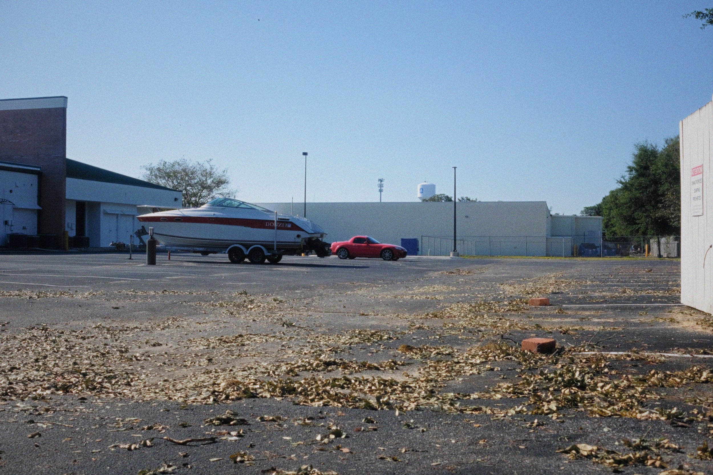 Parking lot with dry leaves scattered on the ground, a white boat on a trailer, and a red sports car parked nearby. Buildings and trees in the background under a clear blue sky.