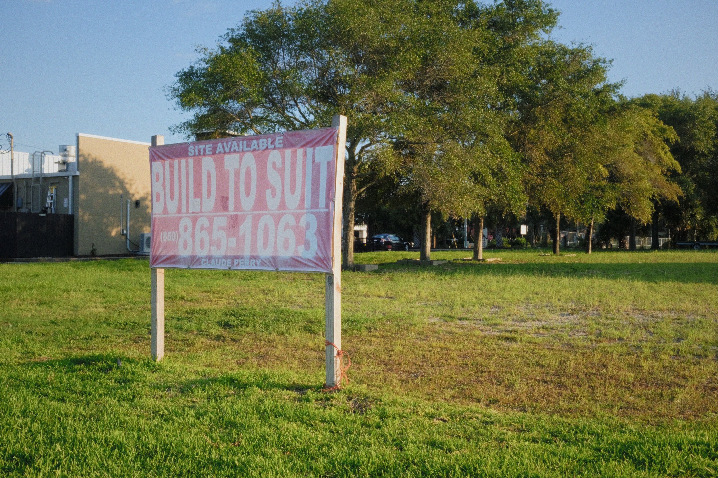 A pink and white sign on a grassy field advertising a new construction site, with the text 'SITE AVAILABLE', 'BUILD TO SUIT', and a phone number. There are trees and a building in the background under a clear blue sky.