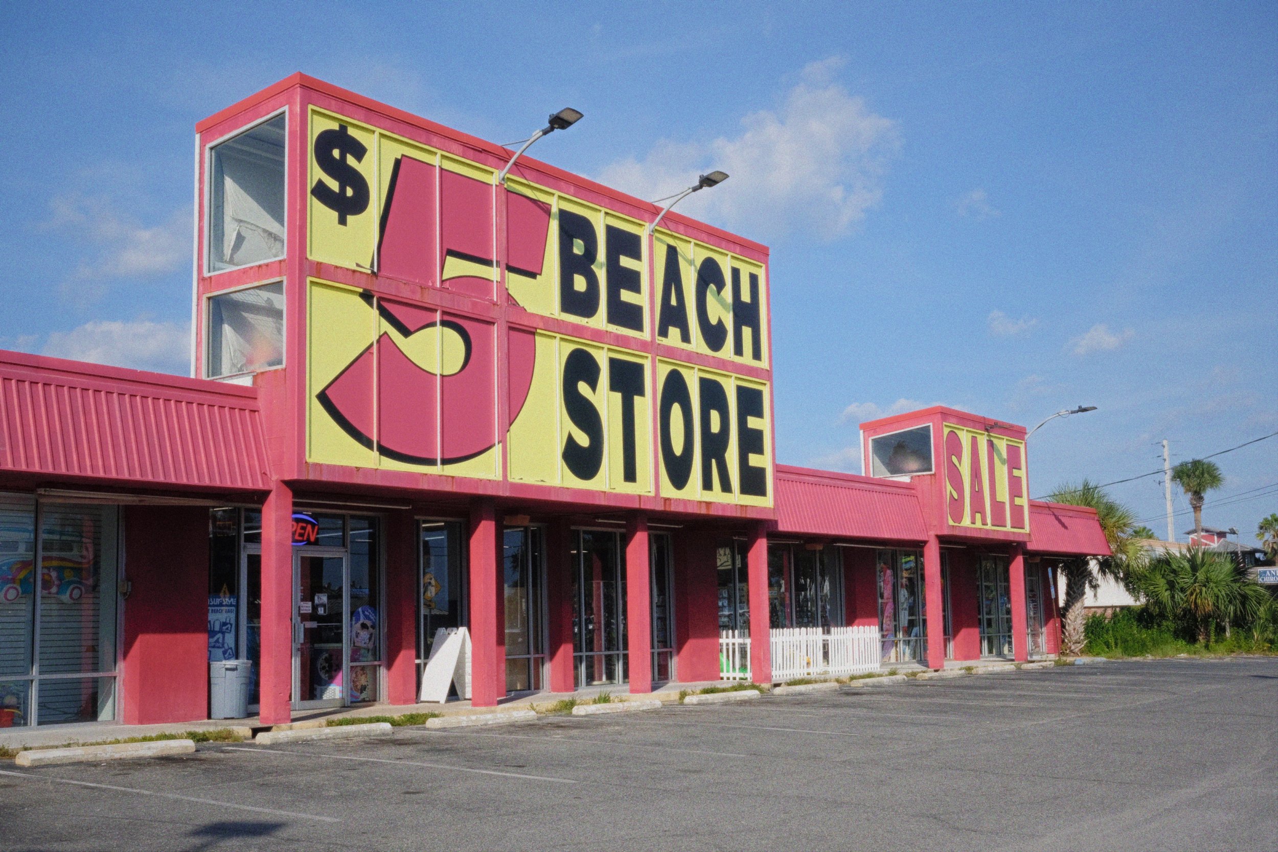 A pink store with large sign reading ' $5 Beach Store' and 'Sale', with an illustration of a watermelon slice, set against a blue sky with scattered clouds and palm trees in the background.