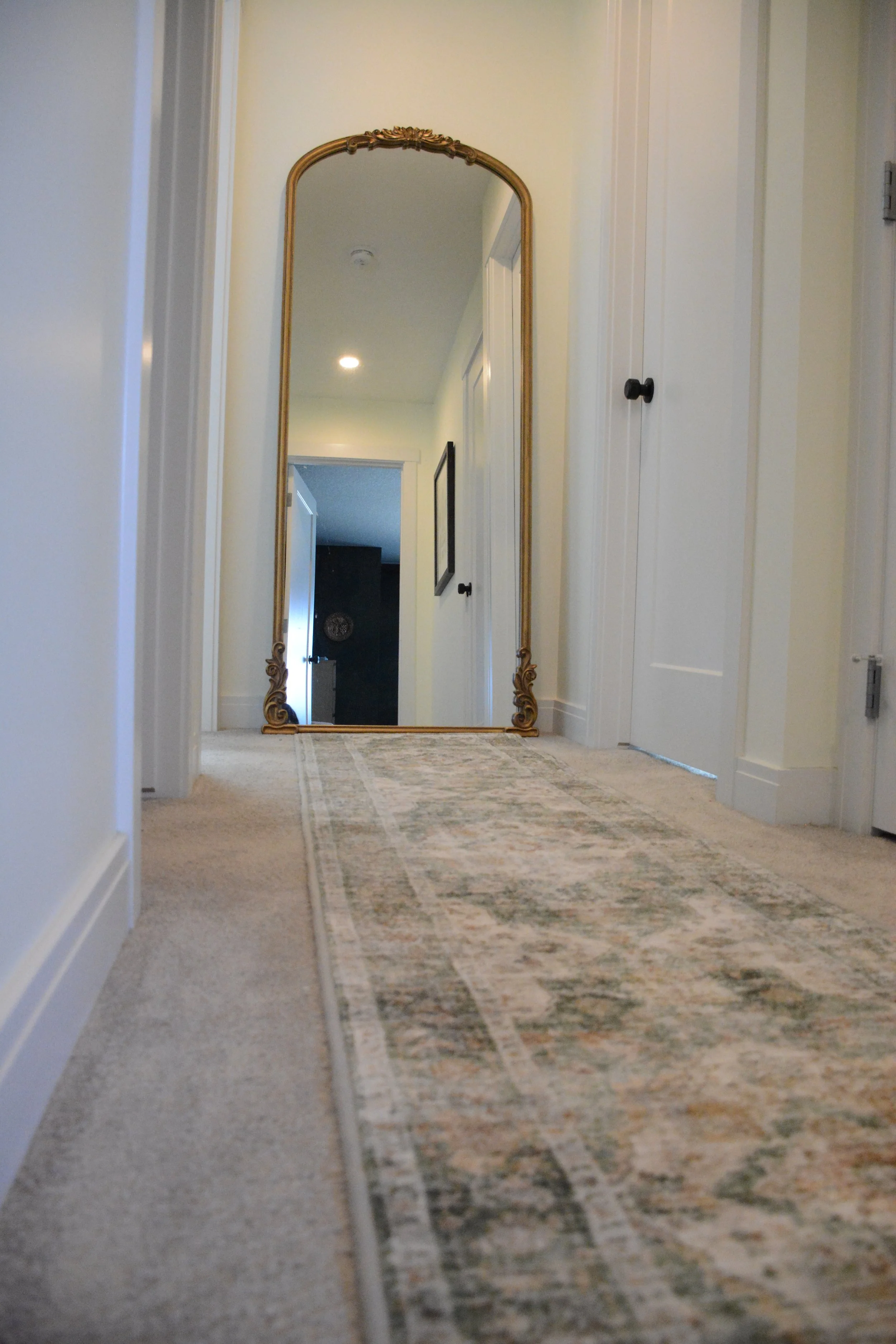 A hallway with a large ornate floor mirror leaning against the wall, a hallway runner rug, and closed white doors on either side.
