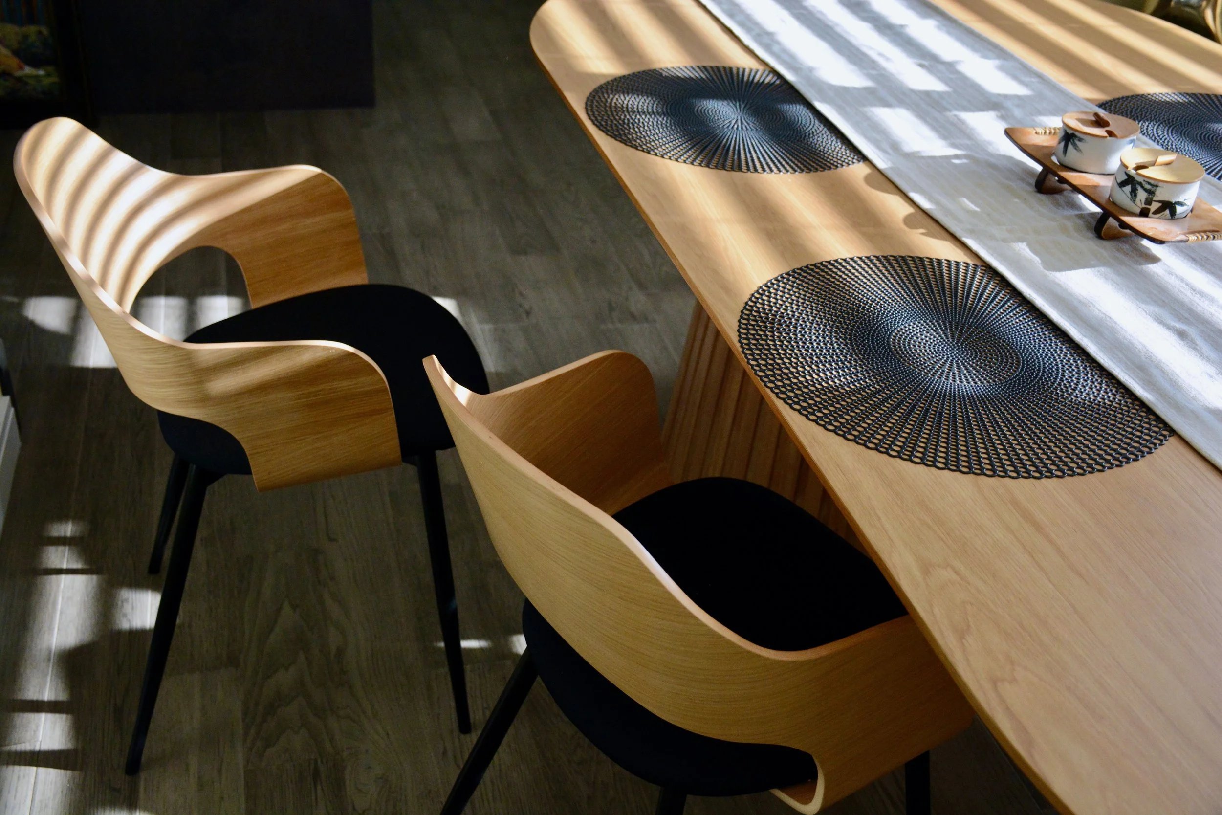 A wooden dining table with black circular placemats, a white table runner, and decorative bowls on a small wooden tray in a room with dark hardwood flooring.