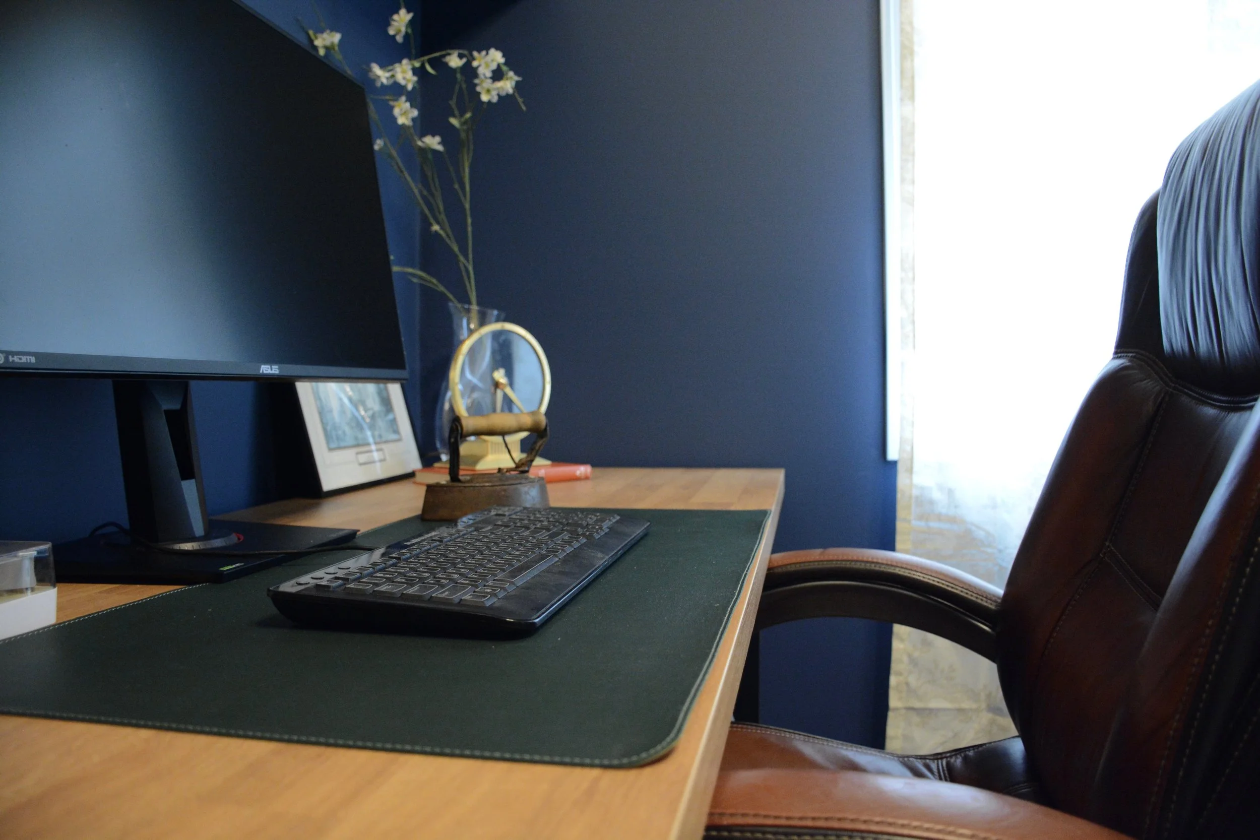 A computer workspace with a monitor, keyboard, and small decorative items on a wooden desk, with a navy blue wall in the background and a window with light coming in.