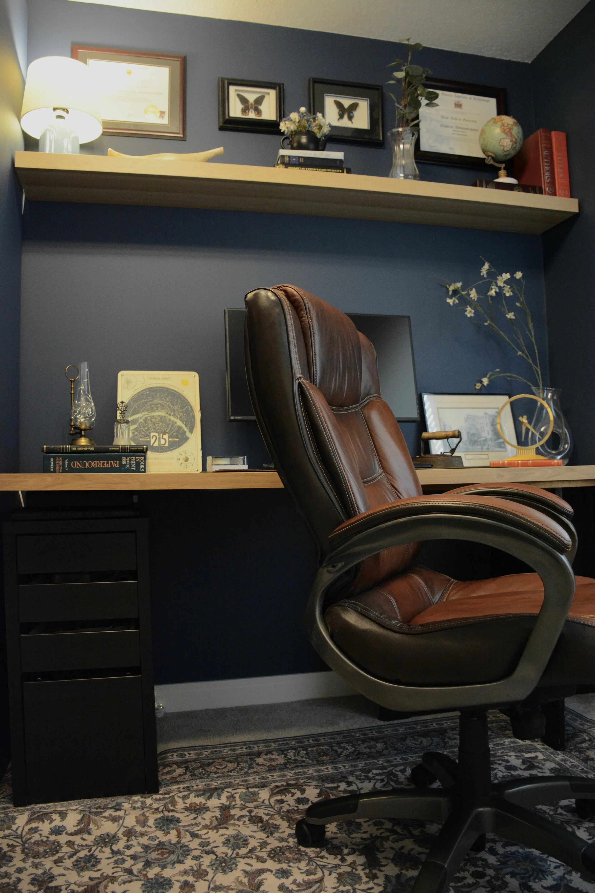A home office with a desk, brown leather office chair, and dark blue walls. The desk has books, a framed picture, and decorative items. A floating shelf above holds framed butterfly pictures, a globe, and plants. A lamp is on the left side of the shelf. The room features a patterned rug and various decorative objects.