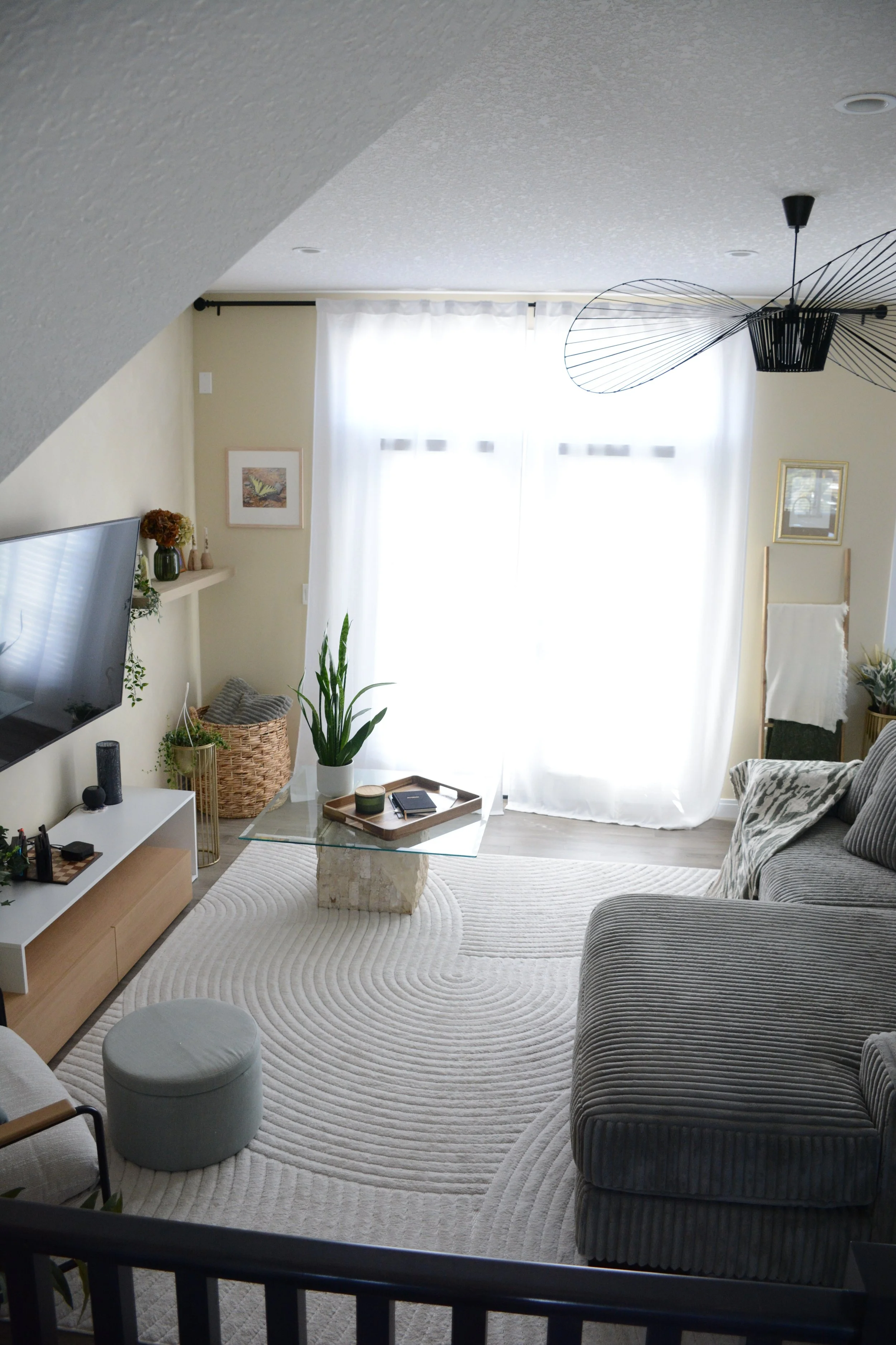 A living room with a gray striped sofa, a glass coffee table on a white rug, a white media console, a flat-screen TV, potted plants, a wicker basket, artwork on the walls, white curtains, a modern black ceiling fan, and a small gray ottoman.