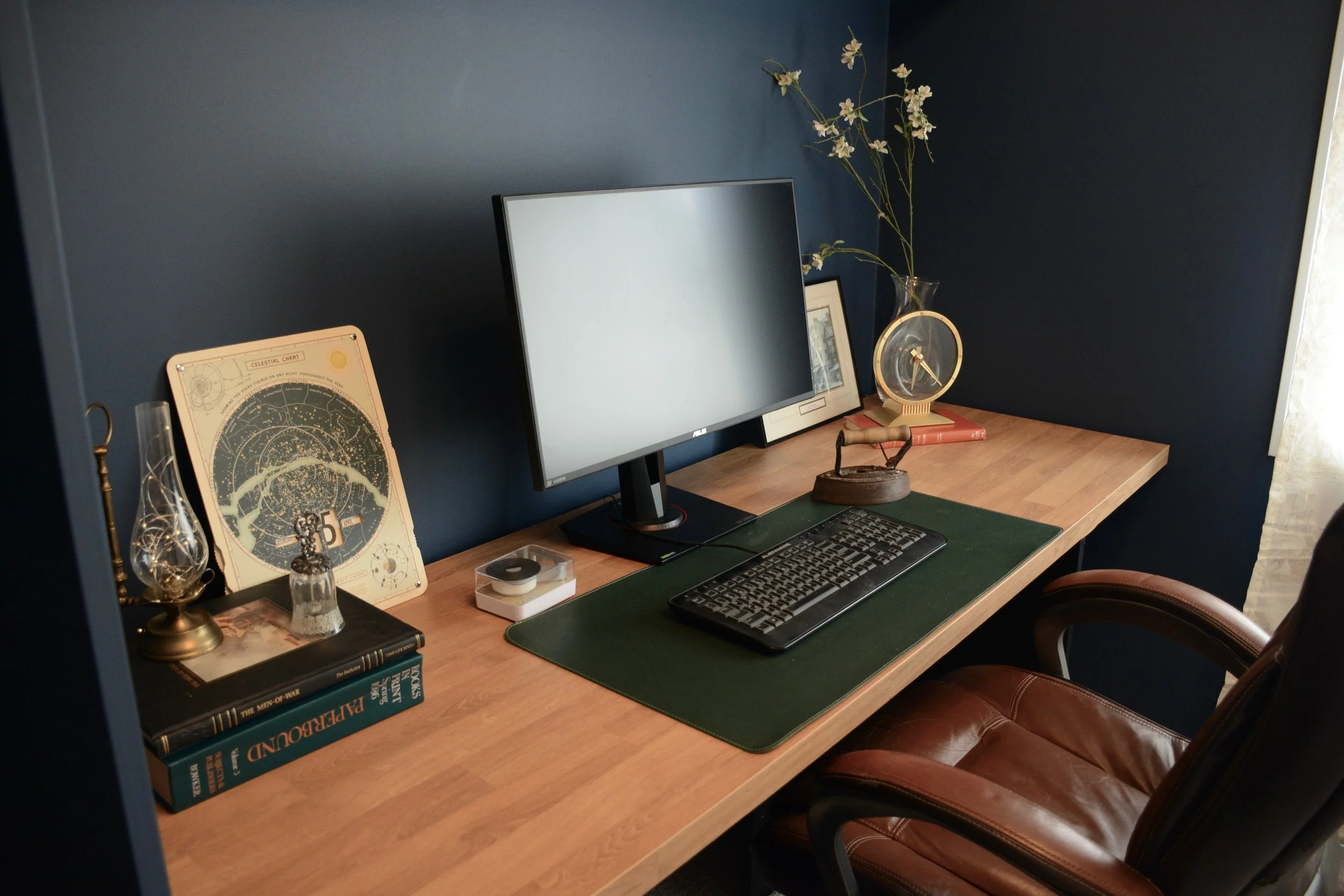 A neat home office desk with an ASUS computer monitor, a keyboard, and a mouse on a green desk mat. Decor items include an astrology chart, a glass light bulb, a decorative bottle, a vintage iron, a clock in a glass vase, and a framed picture. Books are stacked on the left side of the desk, and a brown leather chair is in front of the desk. The wall behind the desk is painted blue.