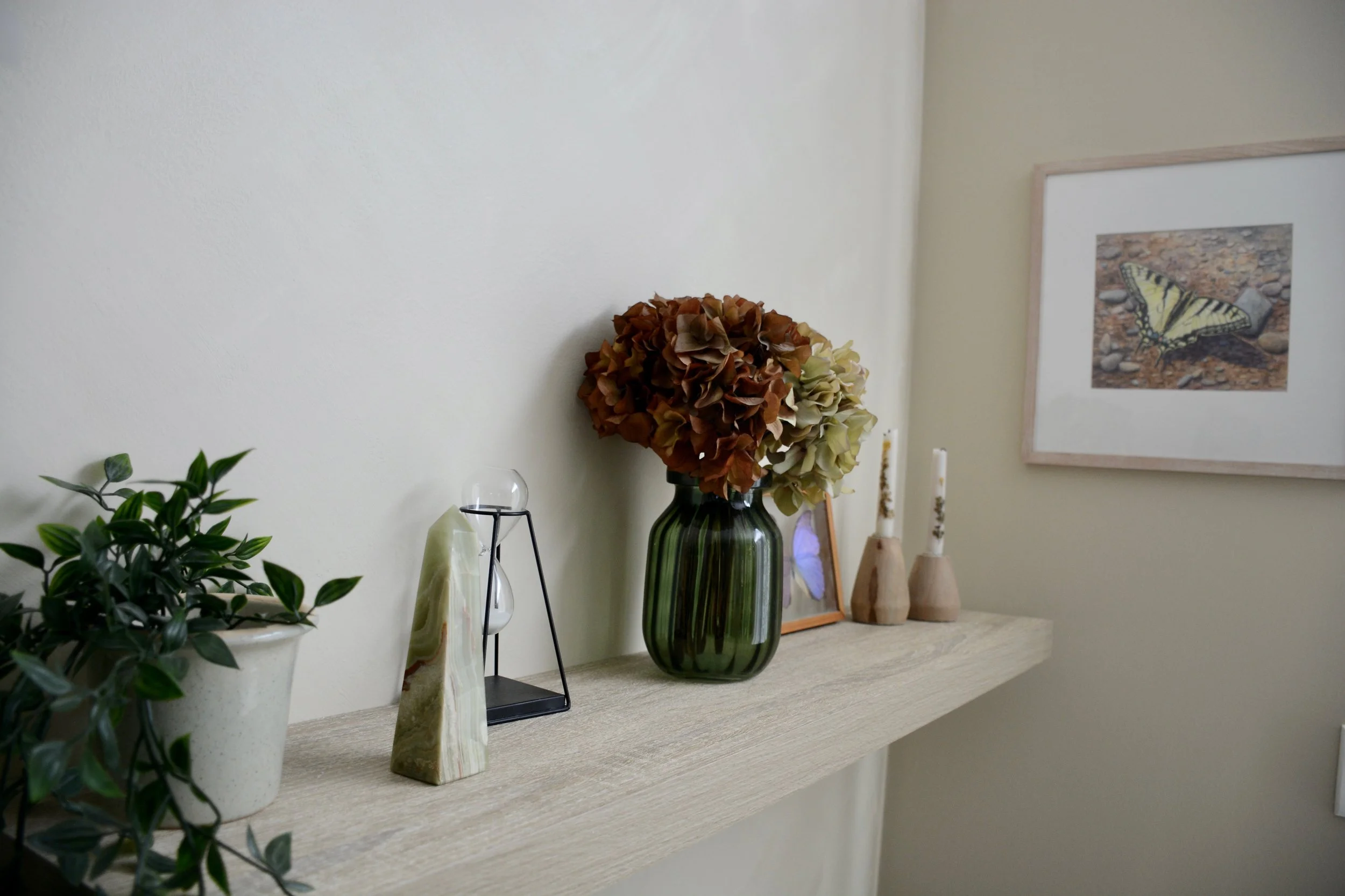 Decorative display on a shelf with a potted plant, a white and green striped stone, decorative objects, a tall green vase with dried hydrangeas, framed artwork of a butterfly, and three wooden candles.