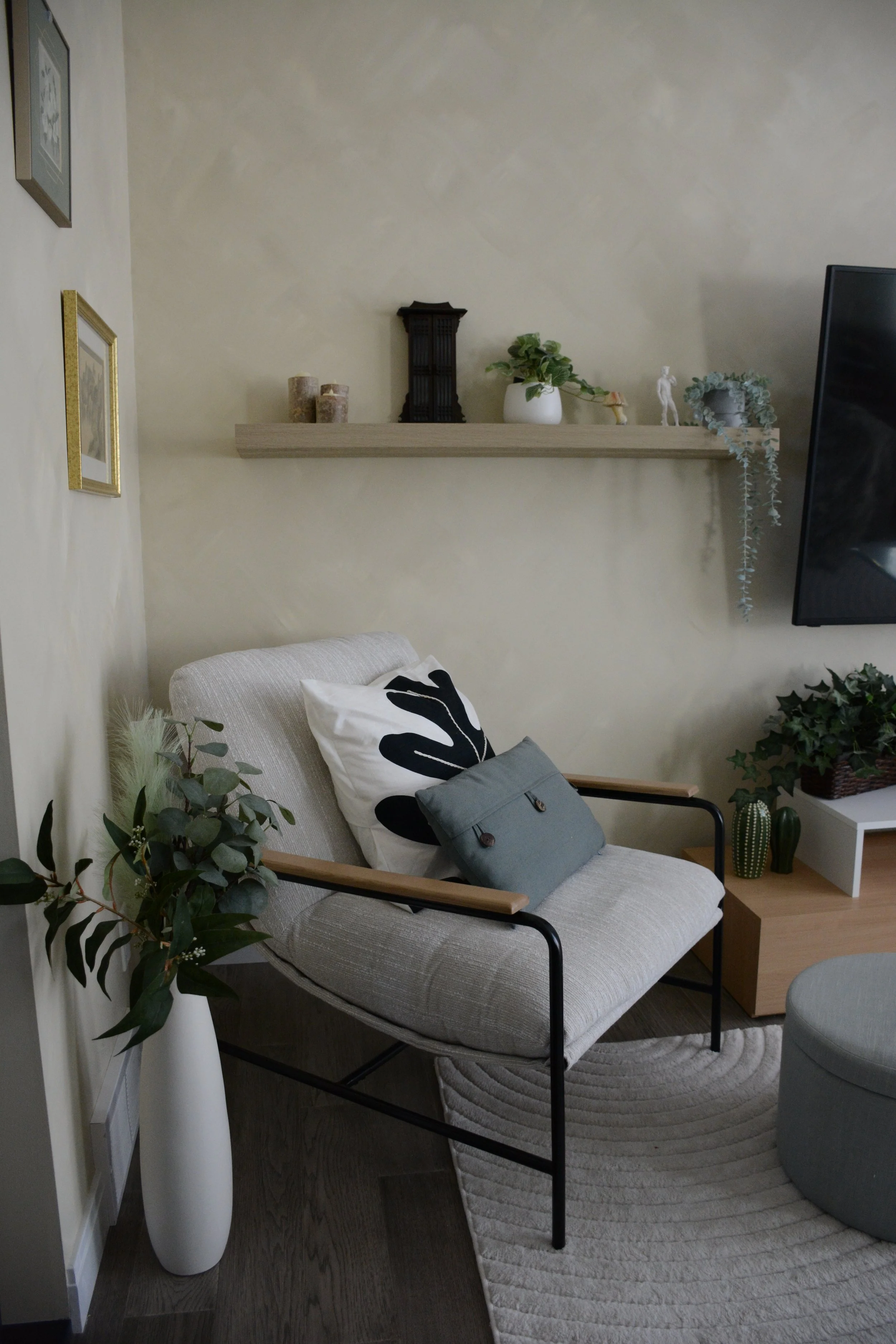 A cozy living room corner with a beige armchair that has wooden armrests, a black and white leaf patterned pillow, and a small gray pillow. Next to the chair is a white vase with green foliage, and a white and beige rug on dark wood flooring. Behind the armchair is a wall-mounted wooden shelf with decorative items including candles, plant, and figurines, and a flat-screen TV on the right.