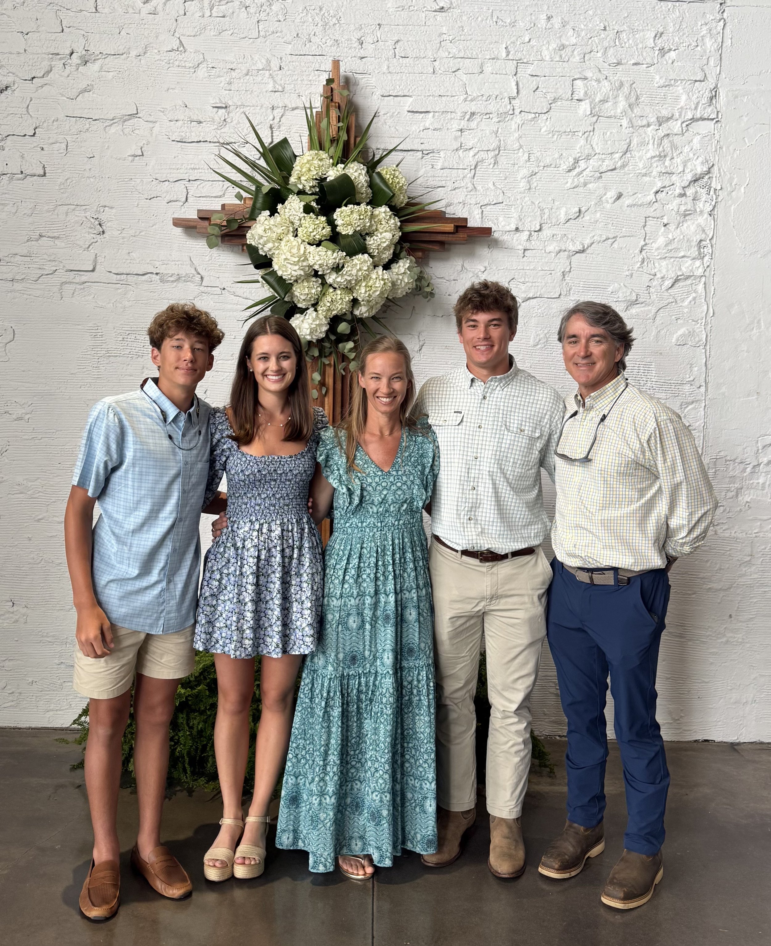 Group of five smiling people standing in front of a white brick wall with a large floral arrangement in the shape of a cross behind them. The group includes three men and two women, dressed casually.