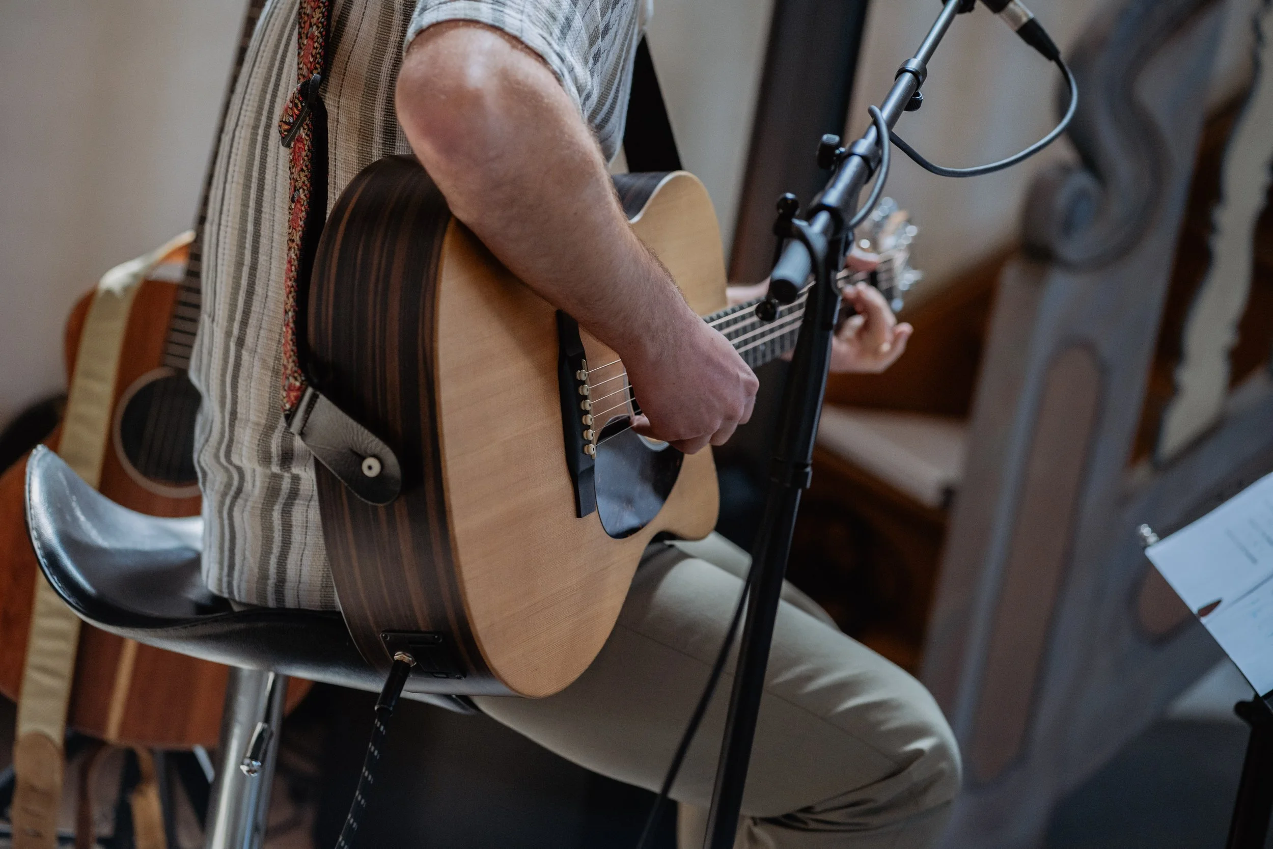 Person playing an acoustic guitar, sitting in a chair.