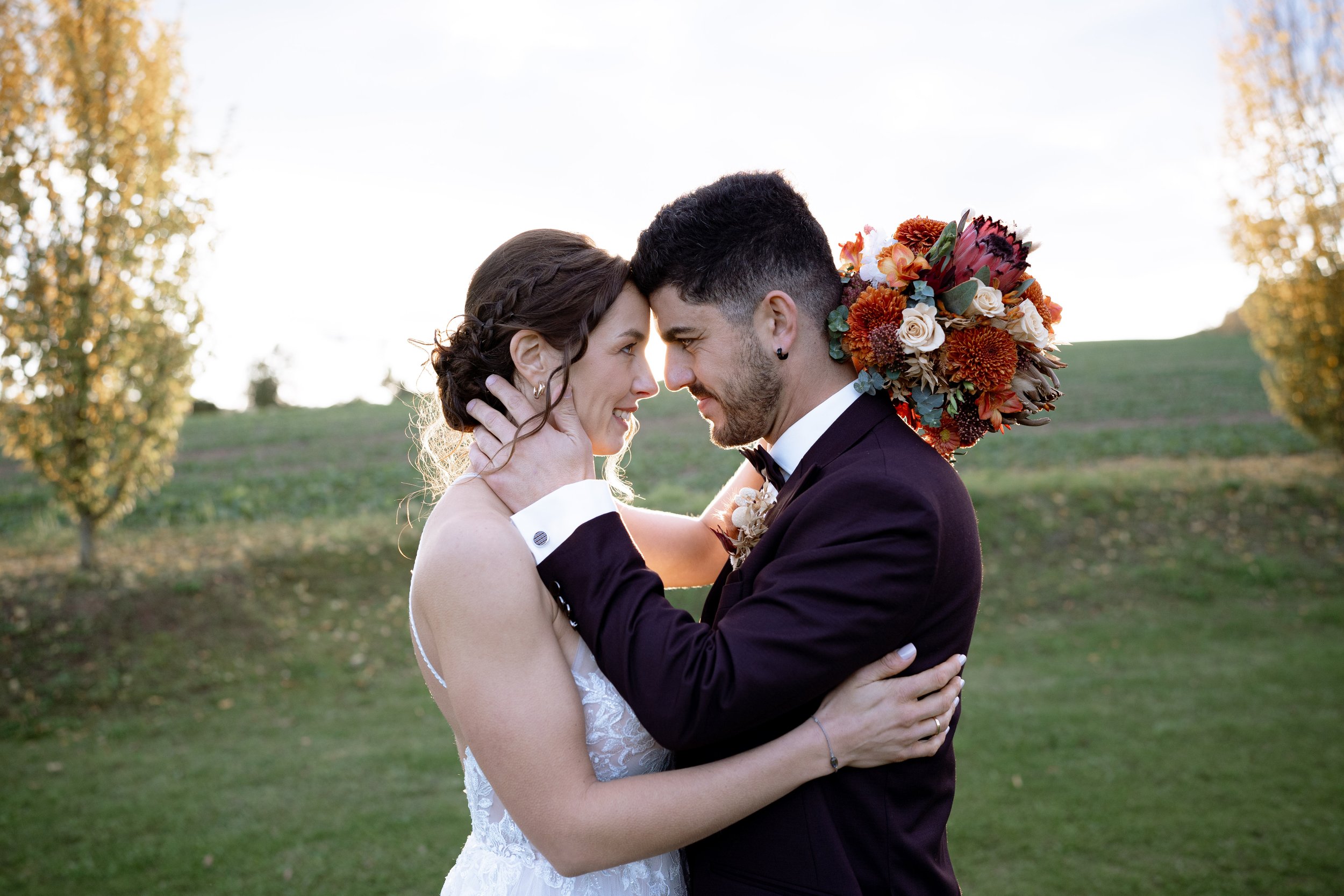 A bride and groom are embracing outdoors with their foreheads touching, smiling at each other. The woman is in a white lace wedding dress and has dark hair, while the man is in a black tuxedo with a decorated flower bouquet on his shoulder. The backg