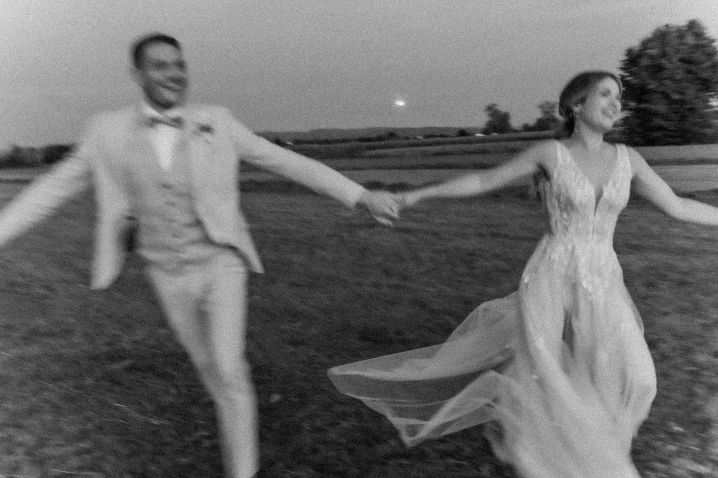 A black and white photo of a smiling couple holding hands and running in a field at dusk, with trees in the background and a bright moon in the sky.