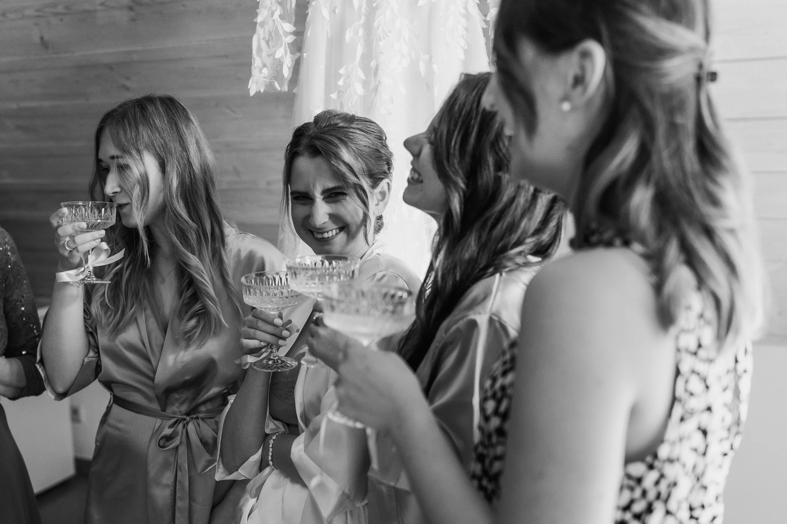 Black and white photo of four women in formal attire, smiling and socializing while holding champagne glasses, at a celebration or party.