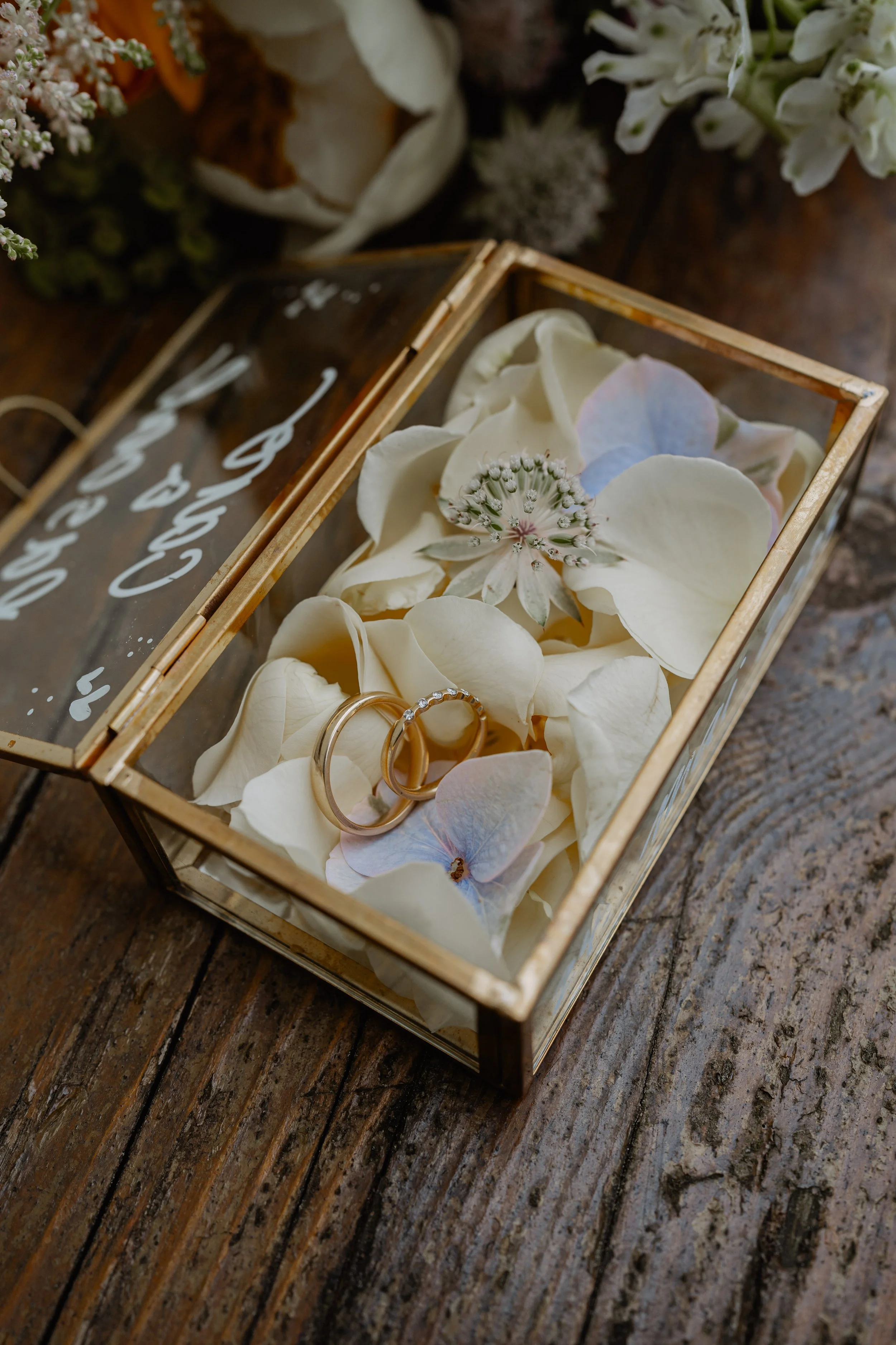 A glass box on a rustic wooden table filled with white and pastel-colored flower petals, with two gold wedding rings resting among the petals.