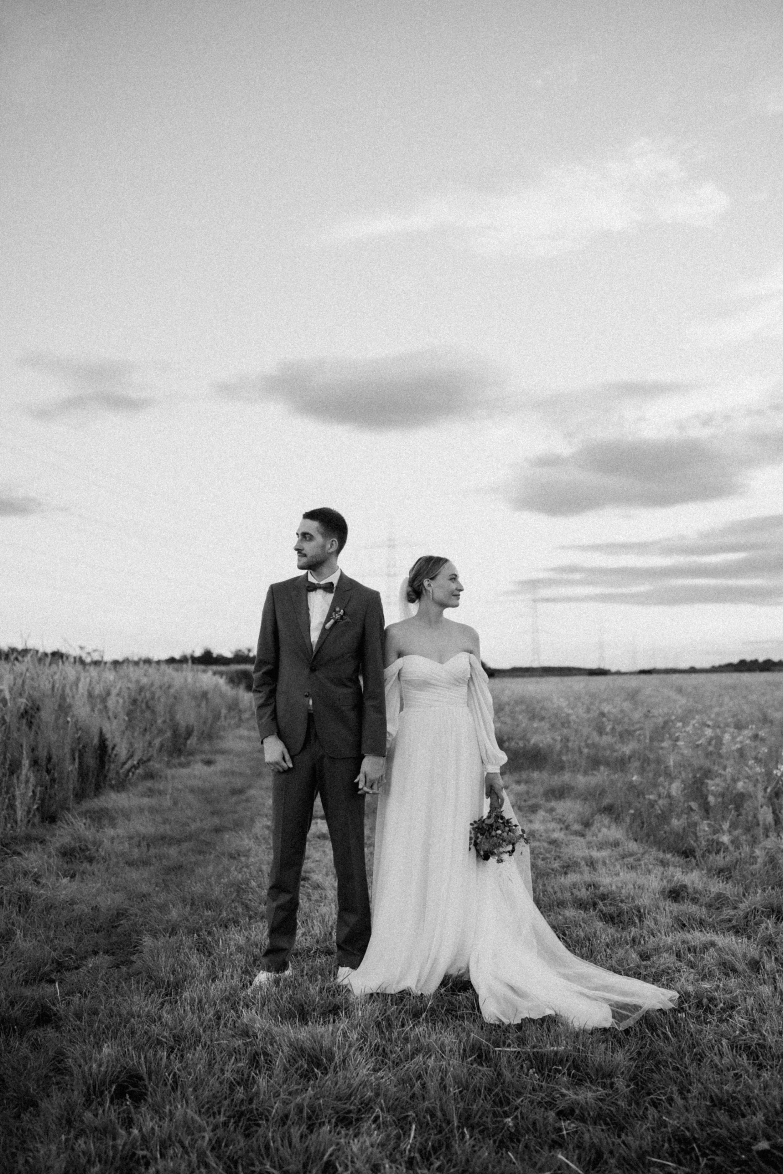 A black and white photograph of a bride and groom standing in a field, holding hands and looking in opposite directions under a cloudy sky.