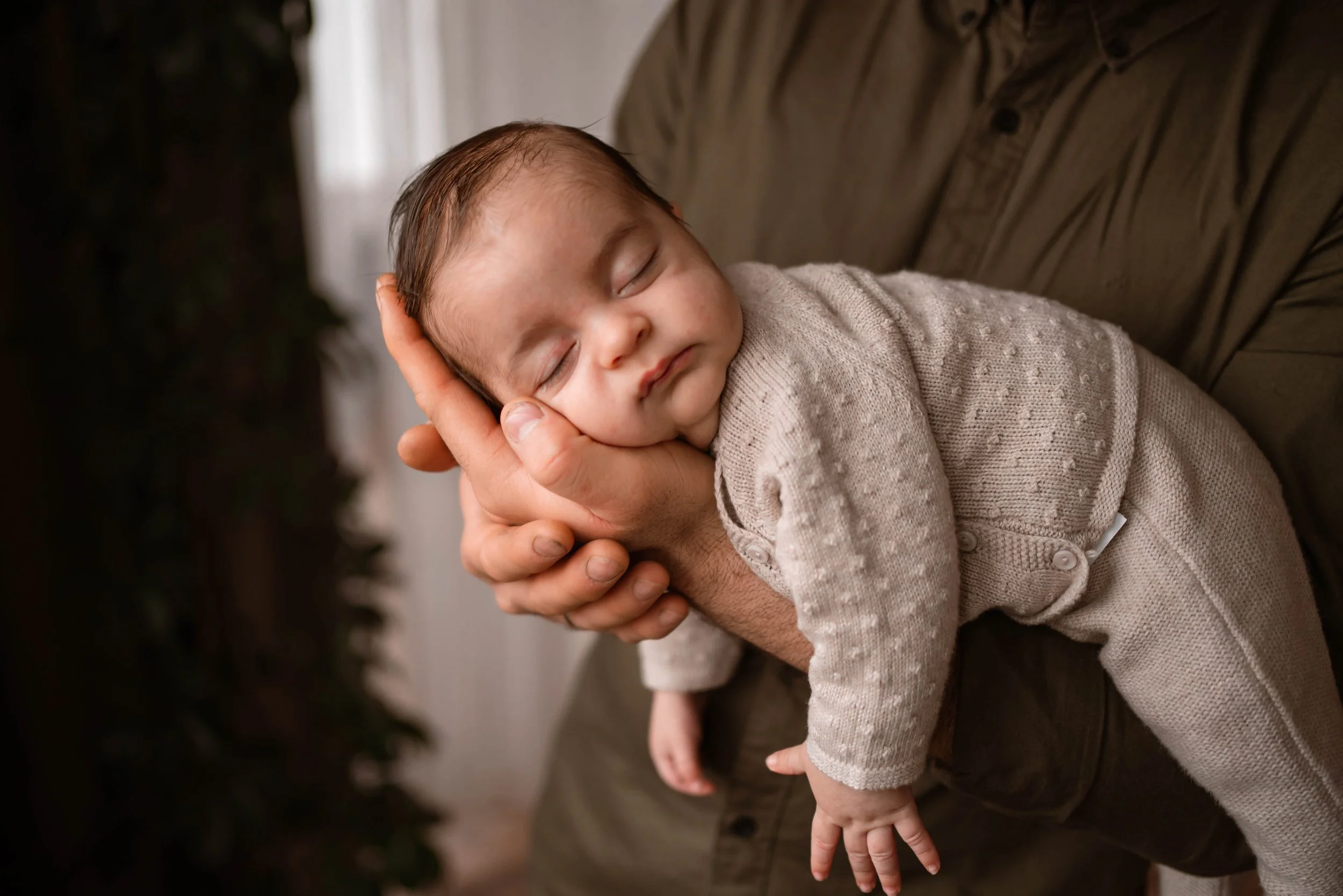 A close-up of a sleeping baby being gently held by an adult with one hand. The baby is dressed in a beige, dotted sweater and is resting on the adult's arm or hand.