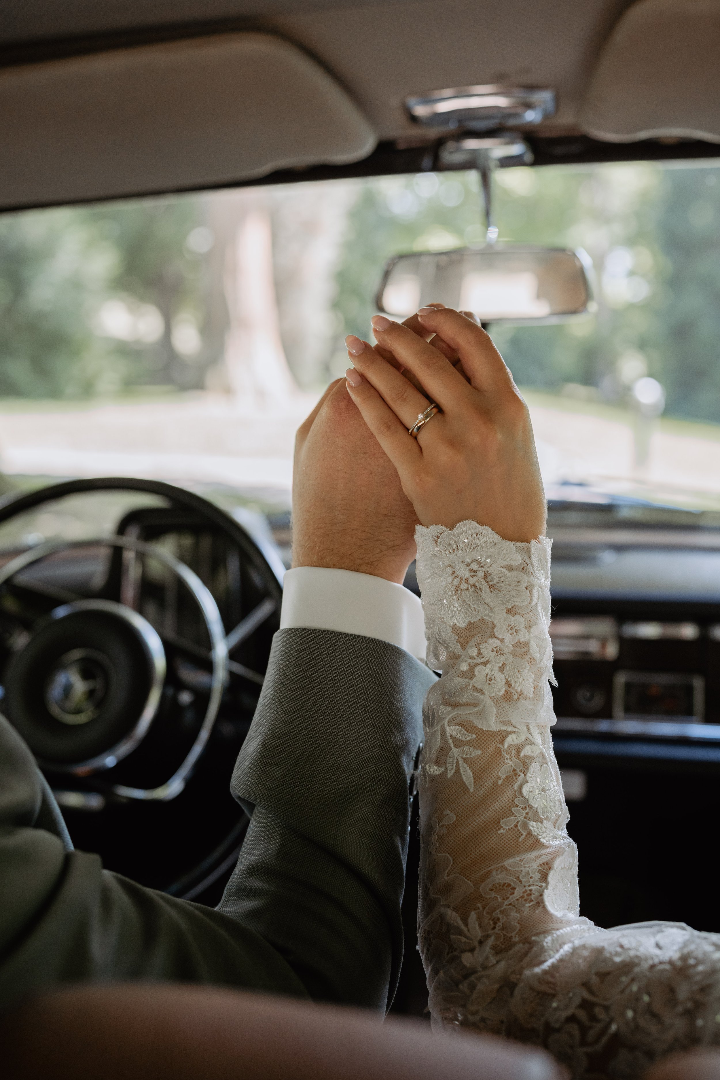 A couple holding hands in a car, with wedding rings visible; the bride is wearing a detailed lace wedding dress, and the groom is in a gray suit. The view is from behind the bride, looking towards the front of the car, with a blurred green outdoor ba