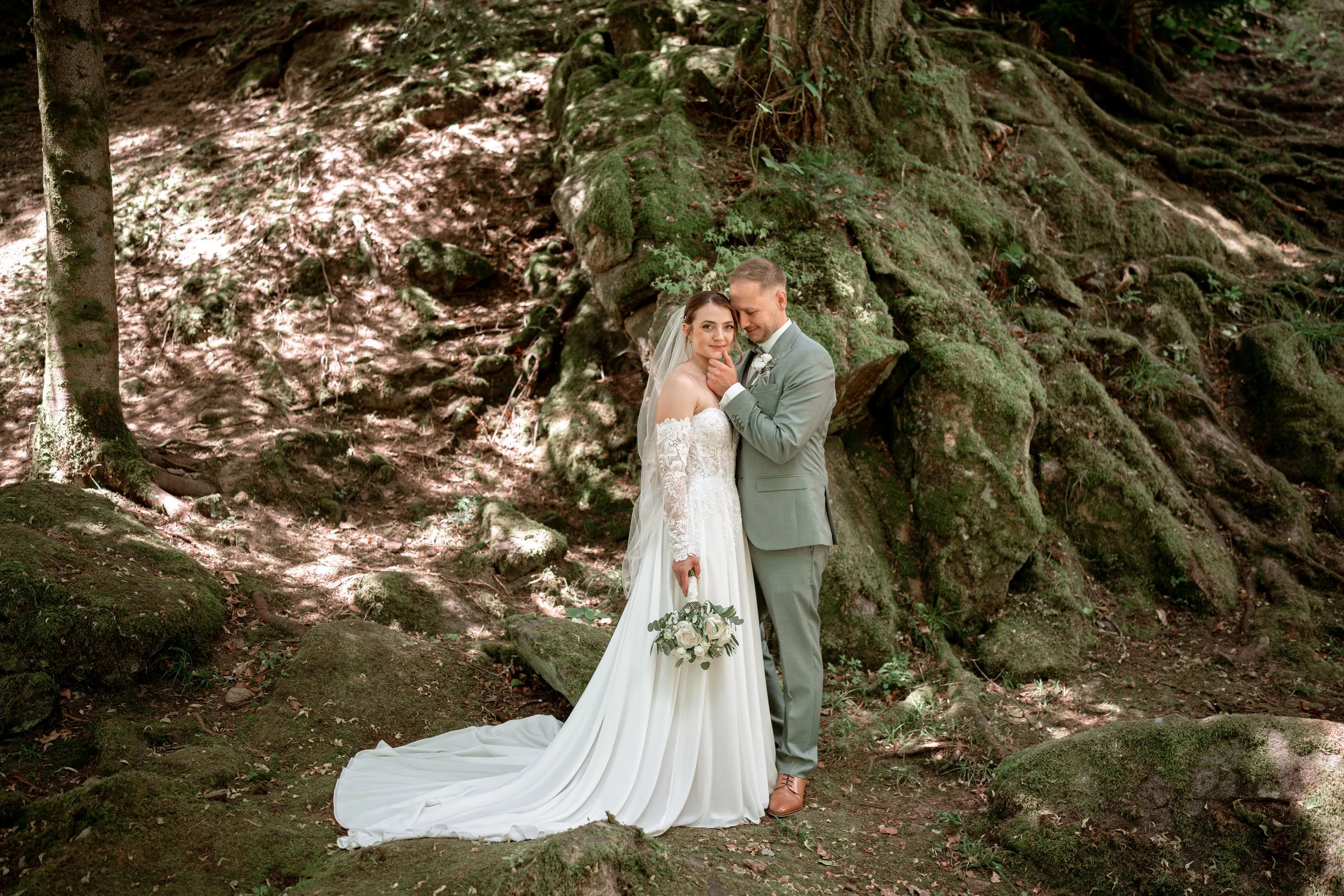 A bride and groom standing close together in a forest, with the bride holding a bouquet of flowers. The bride wears a lace wedding dress and a veil, and the groom is in a gray suit with a white boutonniere. They are smiling softly, surrounded by tree