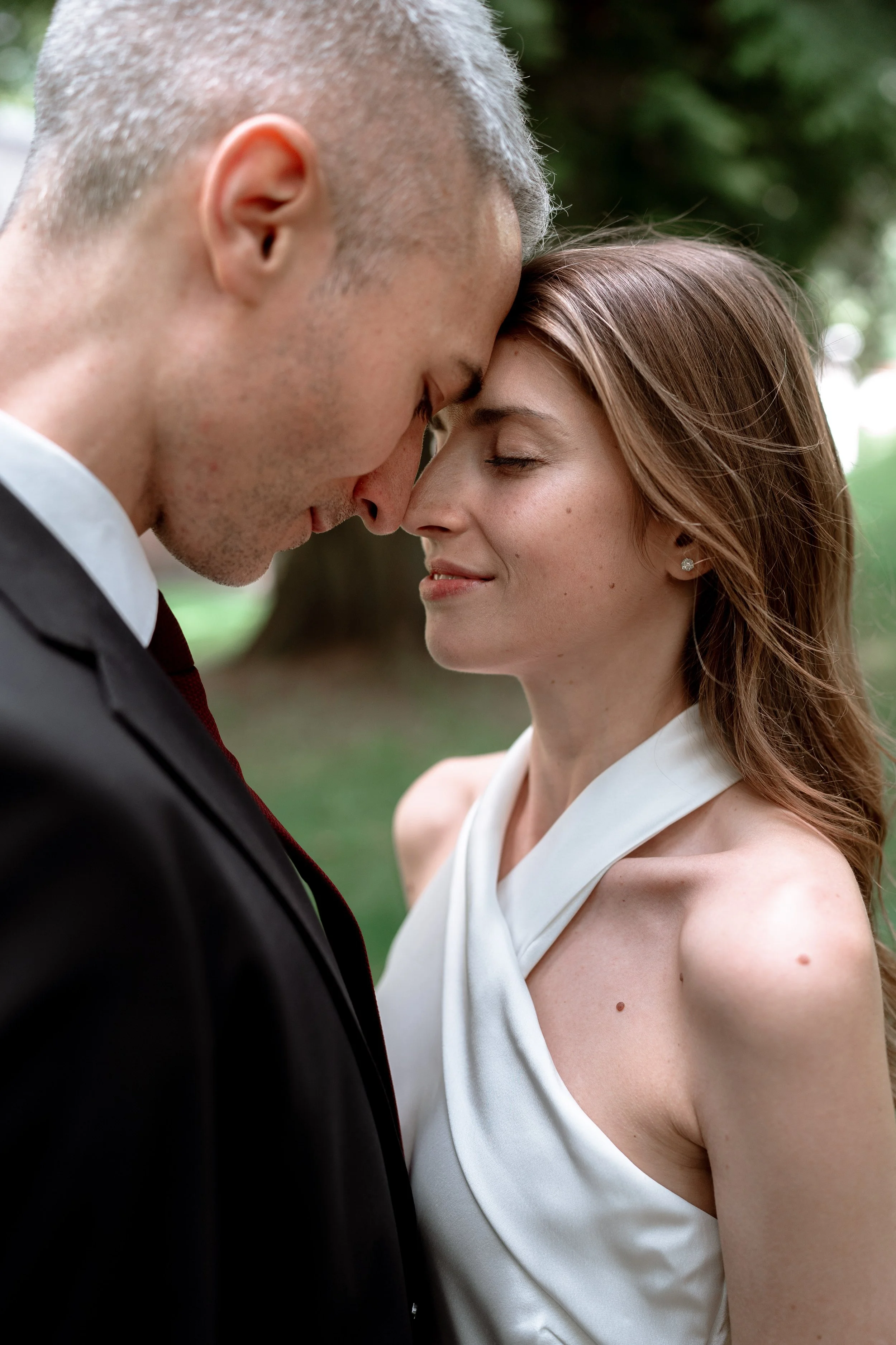 A man in a suit and a woman in a white dress are touching foreheads with their eyes closed, outdoors with blurred greenery in the background.