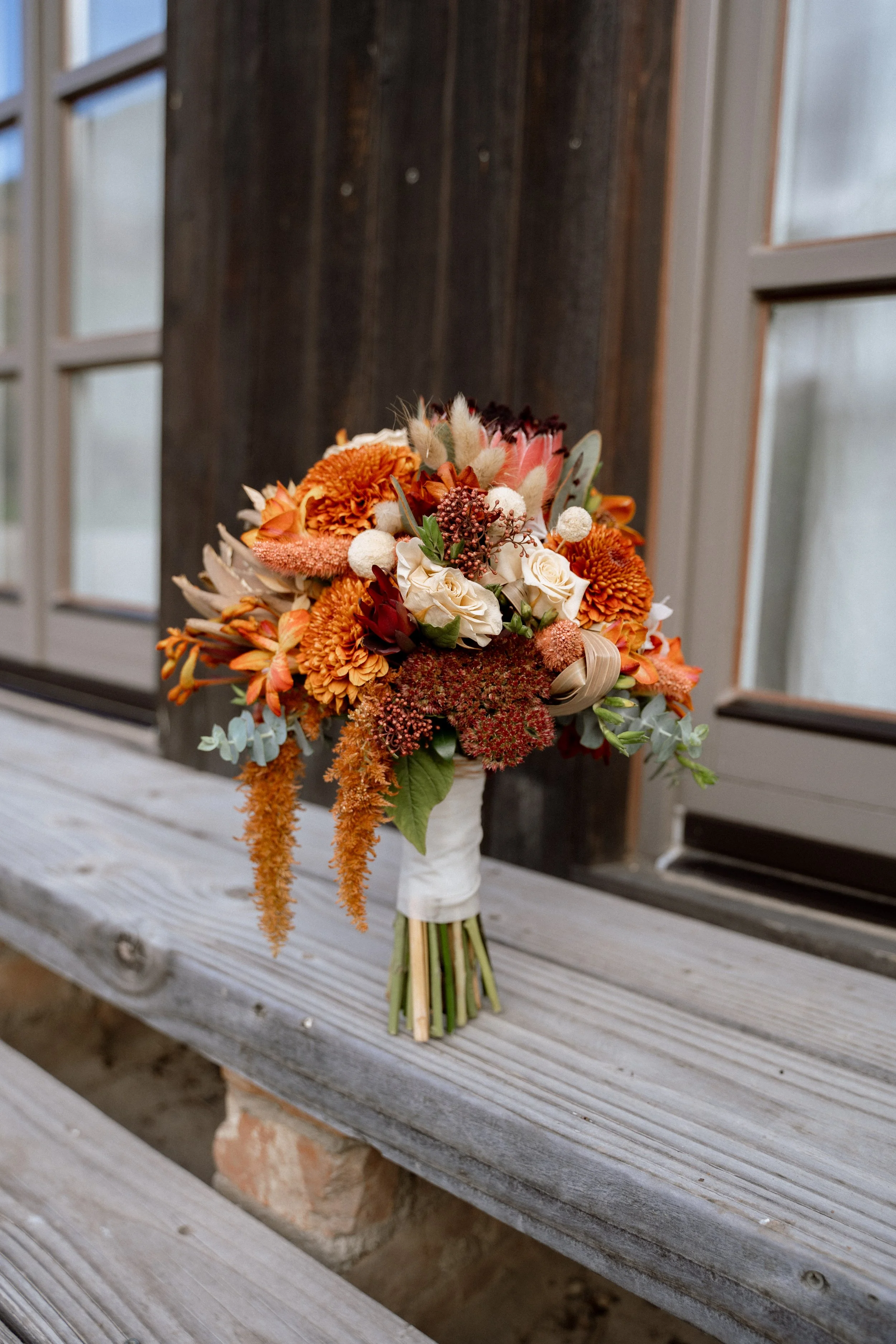 A colorful bouquet of flowers with white, orange, and pink blooms, wrapped with a white ribbon, placed on a wooden bench near a dark brown wall and window frames.