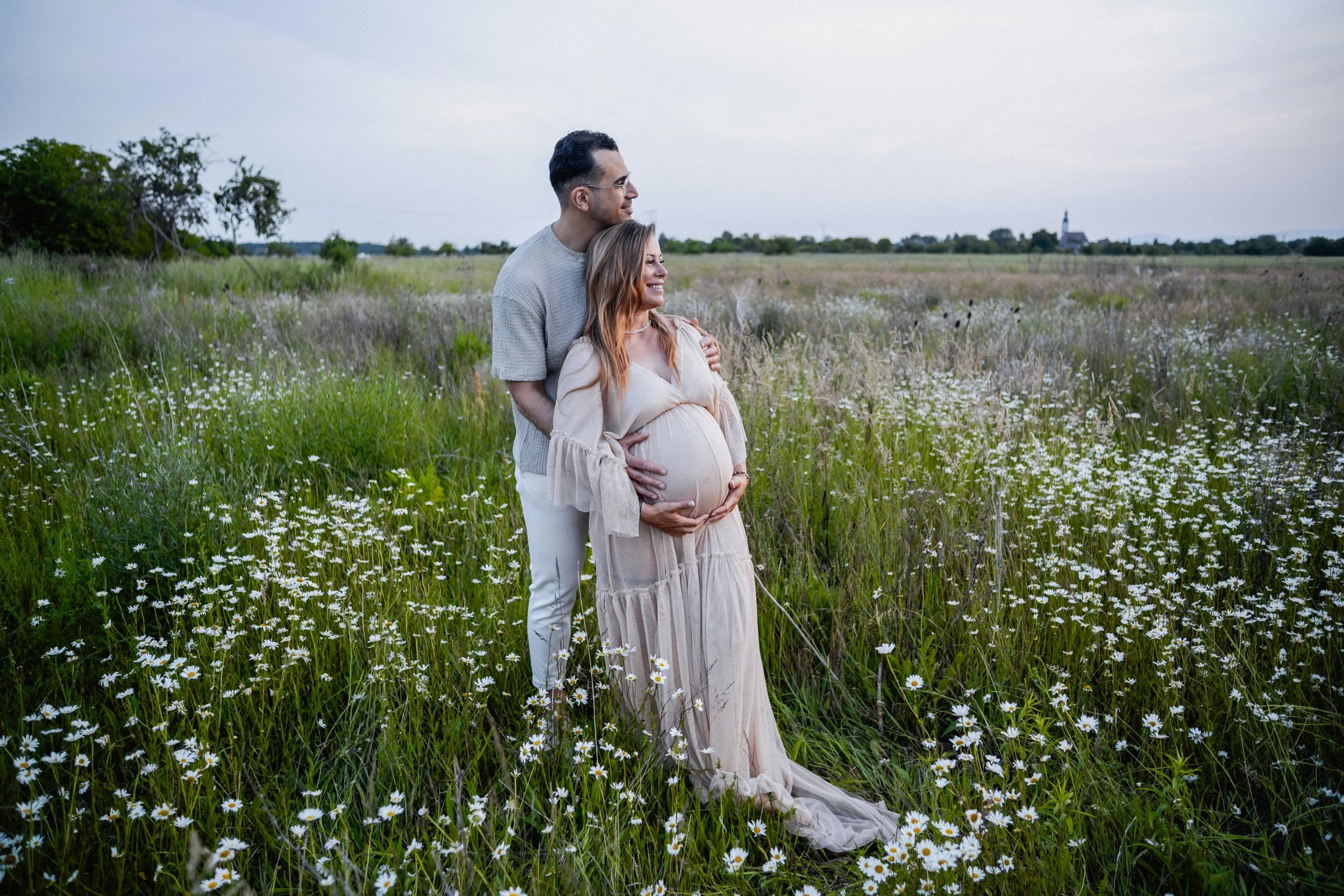 A couple, with the woman pregnant, standing in a field of daisies, with the man embracing her from behind, both smiling, on a cloudy day.