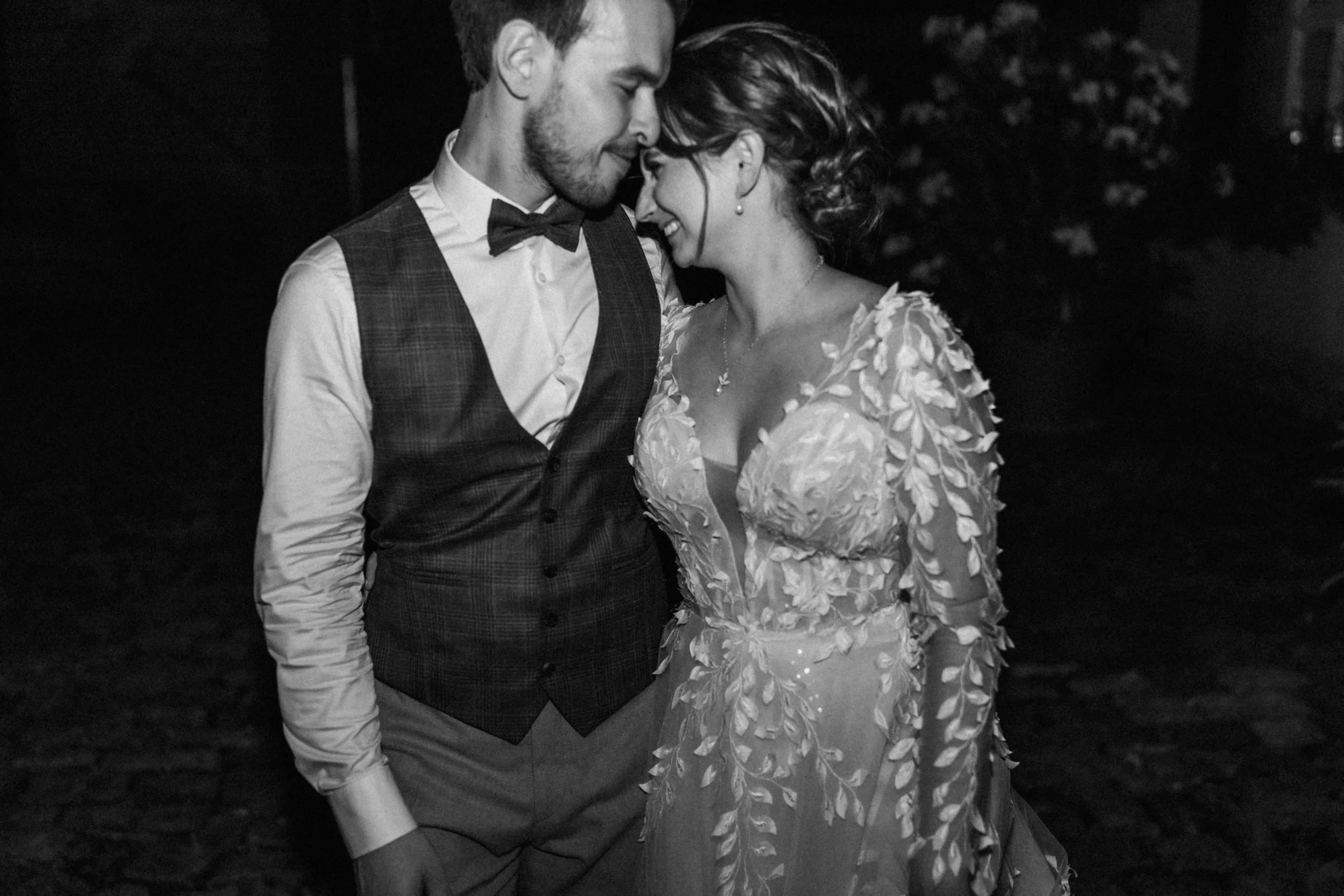 A black and white photo of a couple at a wedding or special event, standing close with their foreheads touching and smiling. The man is wearing a vest, shirt, bow tie, and trousers. The woman is dressed in a gown with floral patterns and has her hair