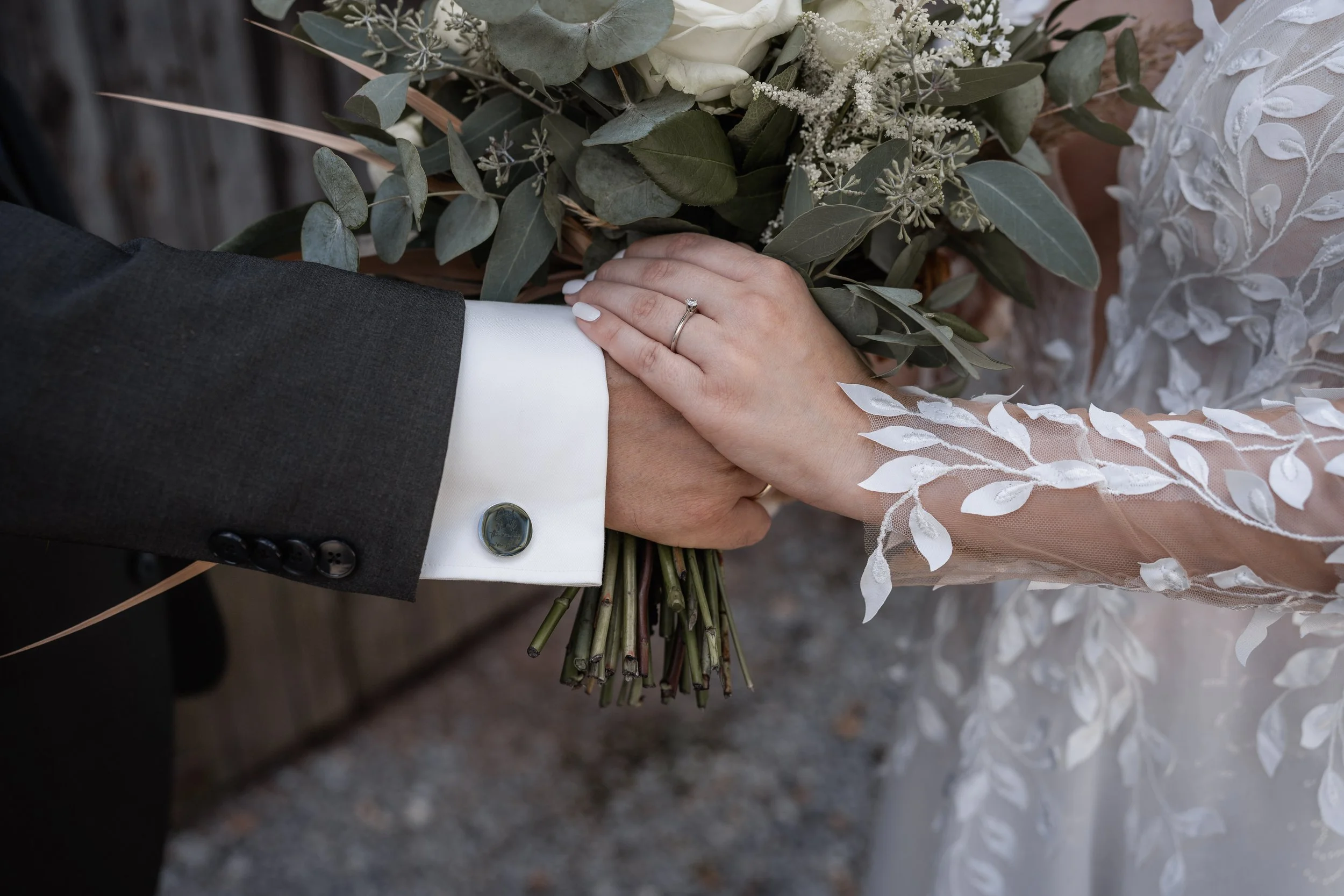 Close-up of a bride and groom holding hands, with the bride's hand resting on the groom's hand. The groom is wearing a dark suit with a white shirt and a buttoned cuff, while the bride's hand, which has a diamond ring, is on top. The bride is wearing