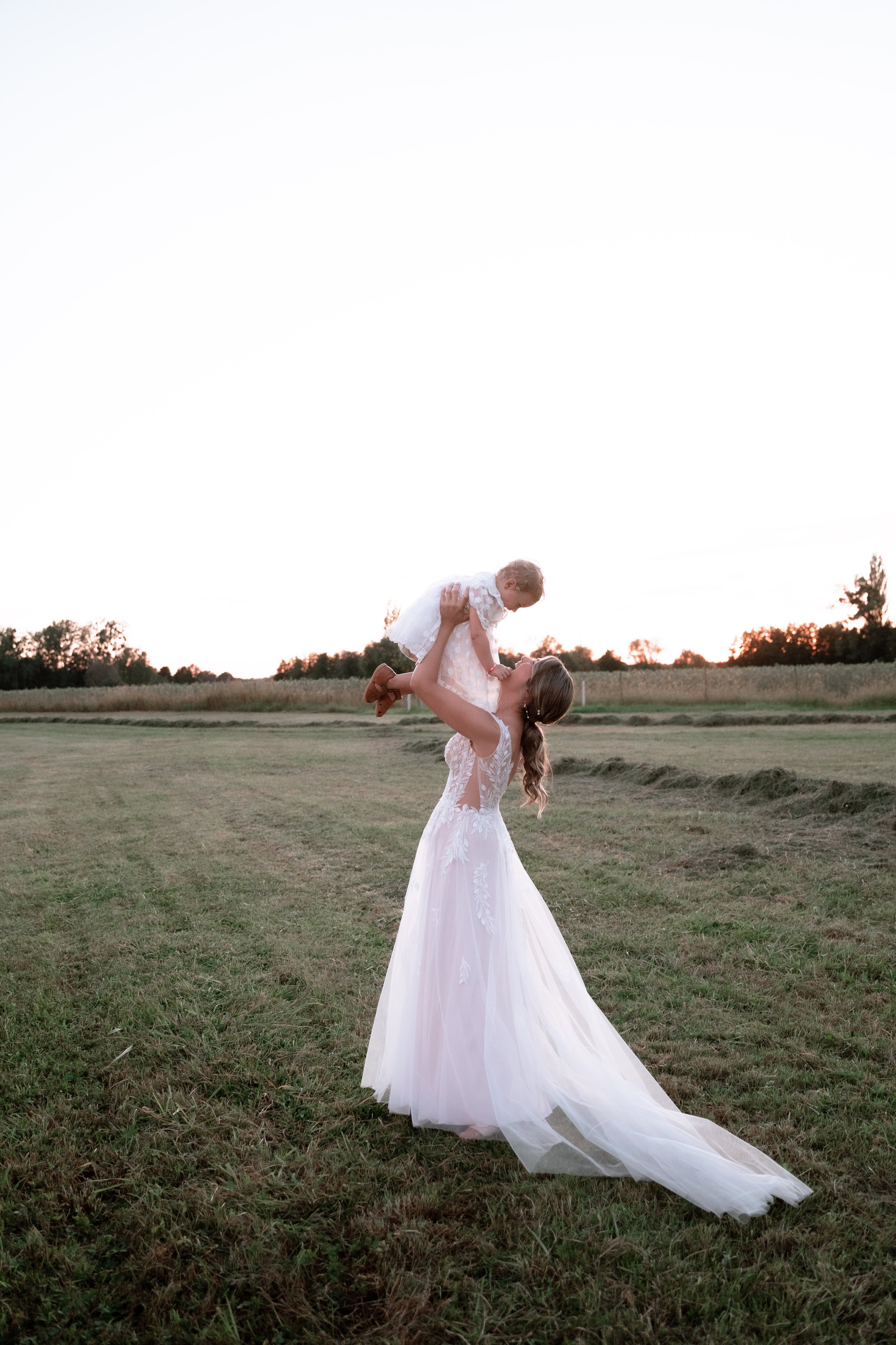 A woman in a white wedding dress holding a young girl wearing a dress in a field during sunset.