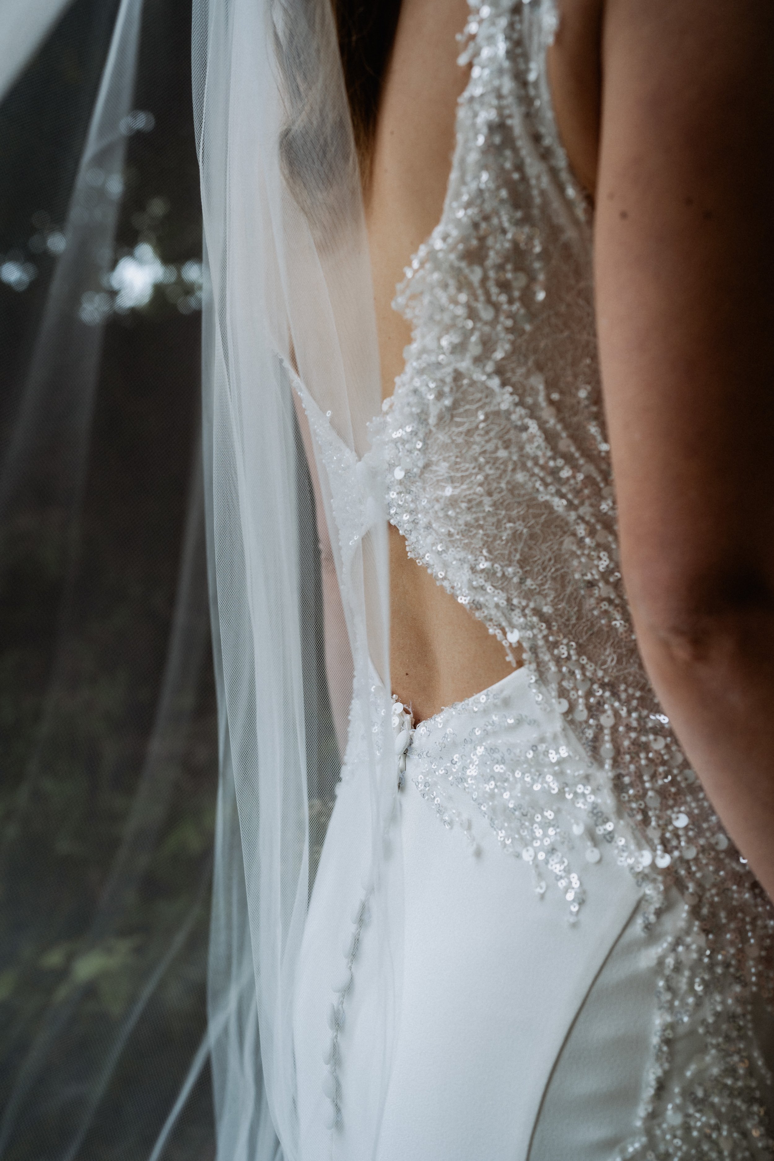 Close-up image of a bride's wedding dress with sequins, viewed from the side through a window, with a veil partially visible.