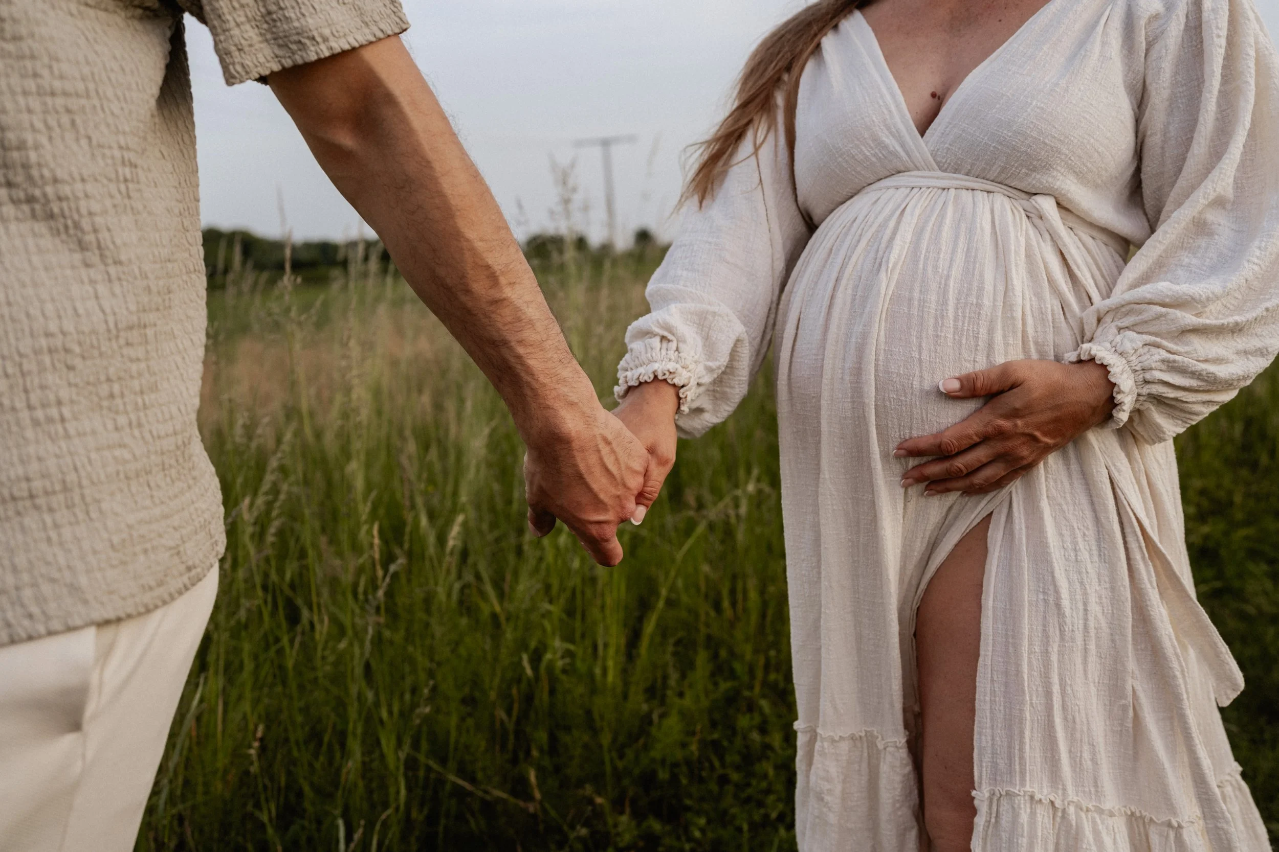 A couple holding hands in a grassy field, with the woman pregnant and wearing a white dress, and the man wearing a beige textured shirt.