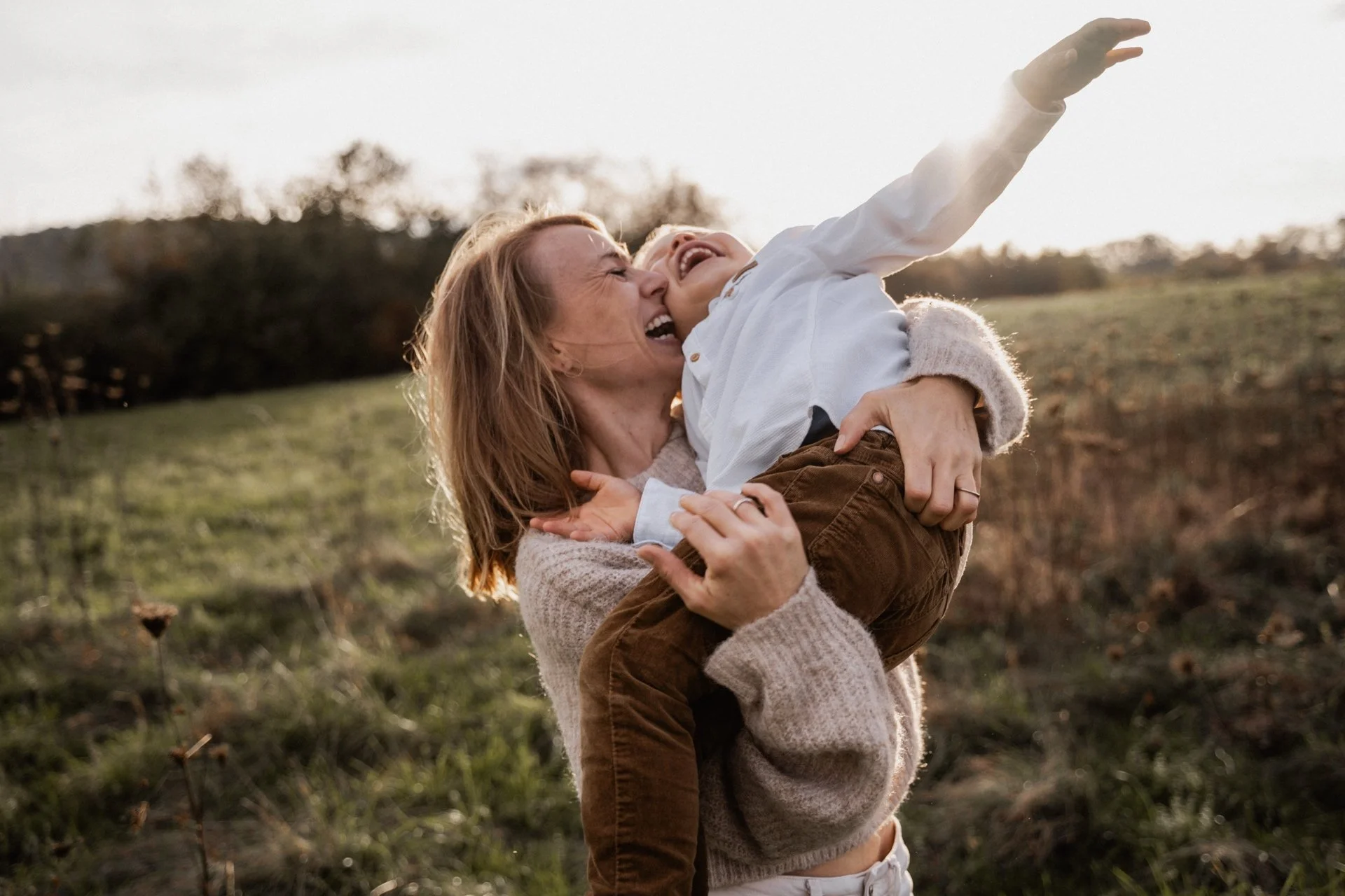 A woman lifting a laughing boy outdoors during sunset, with trees and a grassy field in the background.