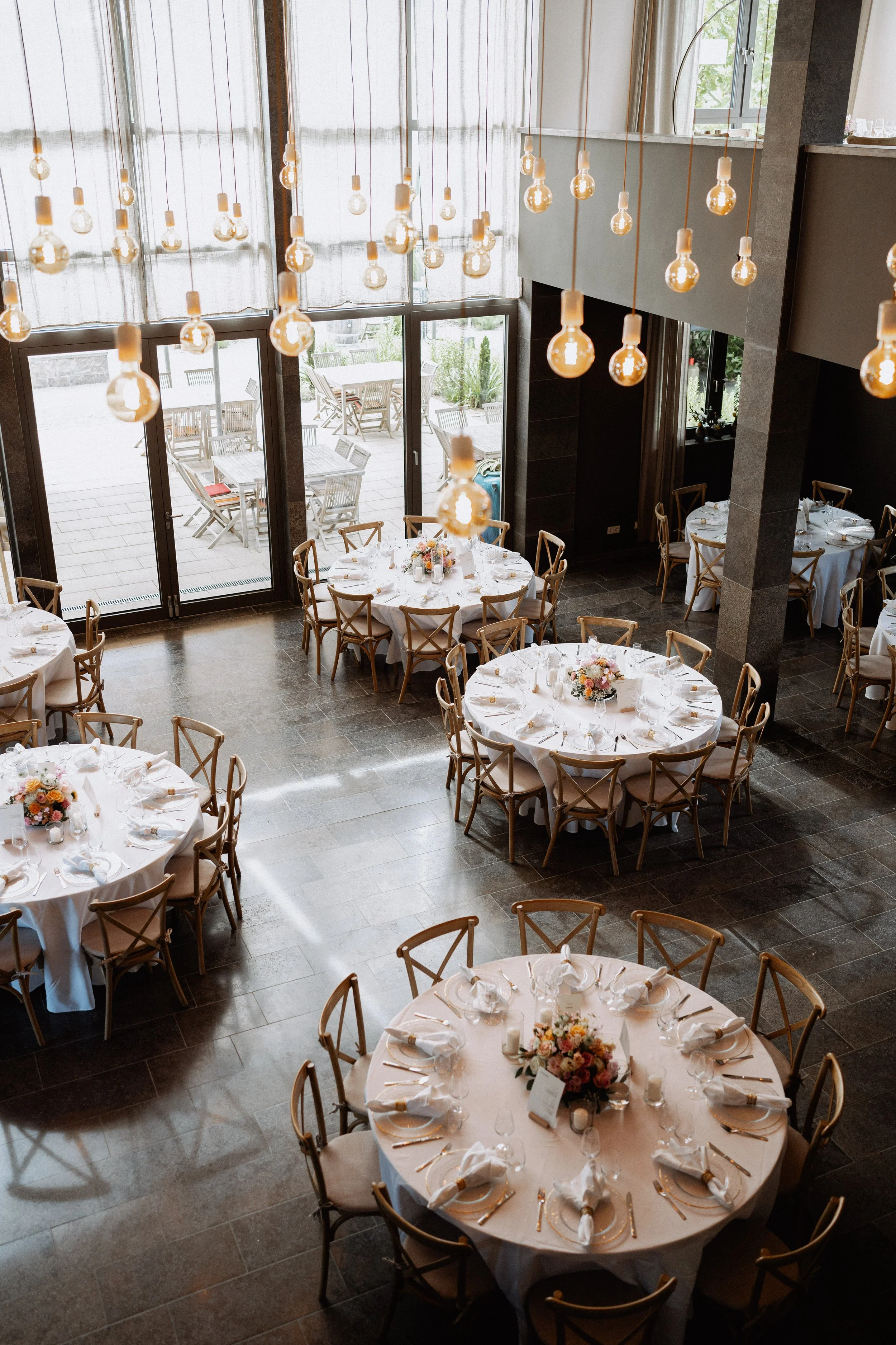 Elegant banquet hall with round tables set for a formal event, decorated with floral centerpieces, white tablecloths, gold cutlery, and napkins, with pendant lights hanging from the ceiling and a large glass door and window showing an outdoor patio a
