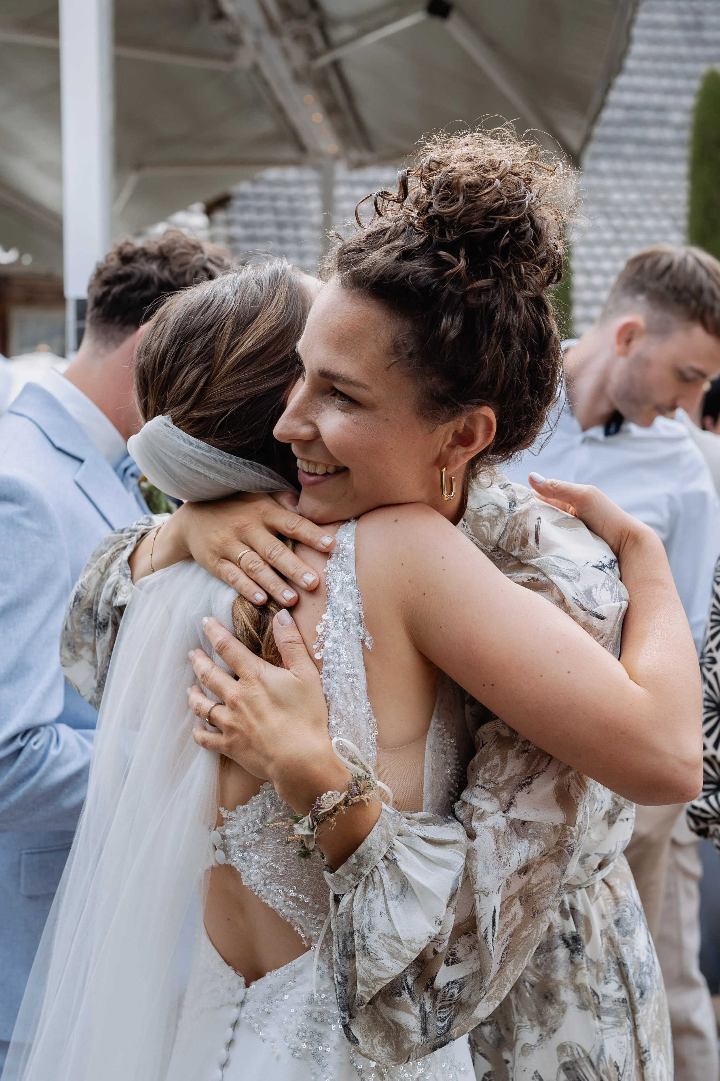 A woman with curly hair and earrings hugging another person wearing a white dress. They are surrounded by other people at an outdoor event, possibly a wedding.