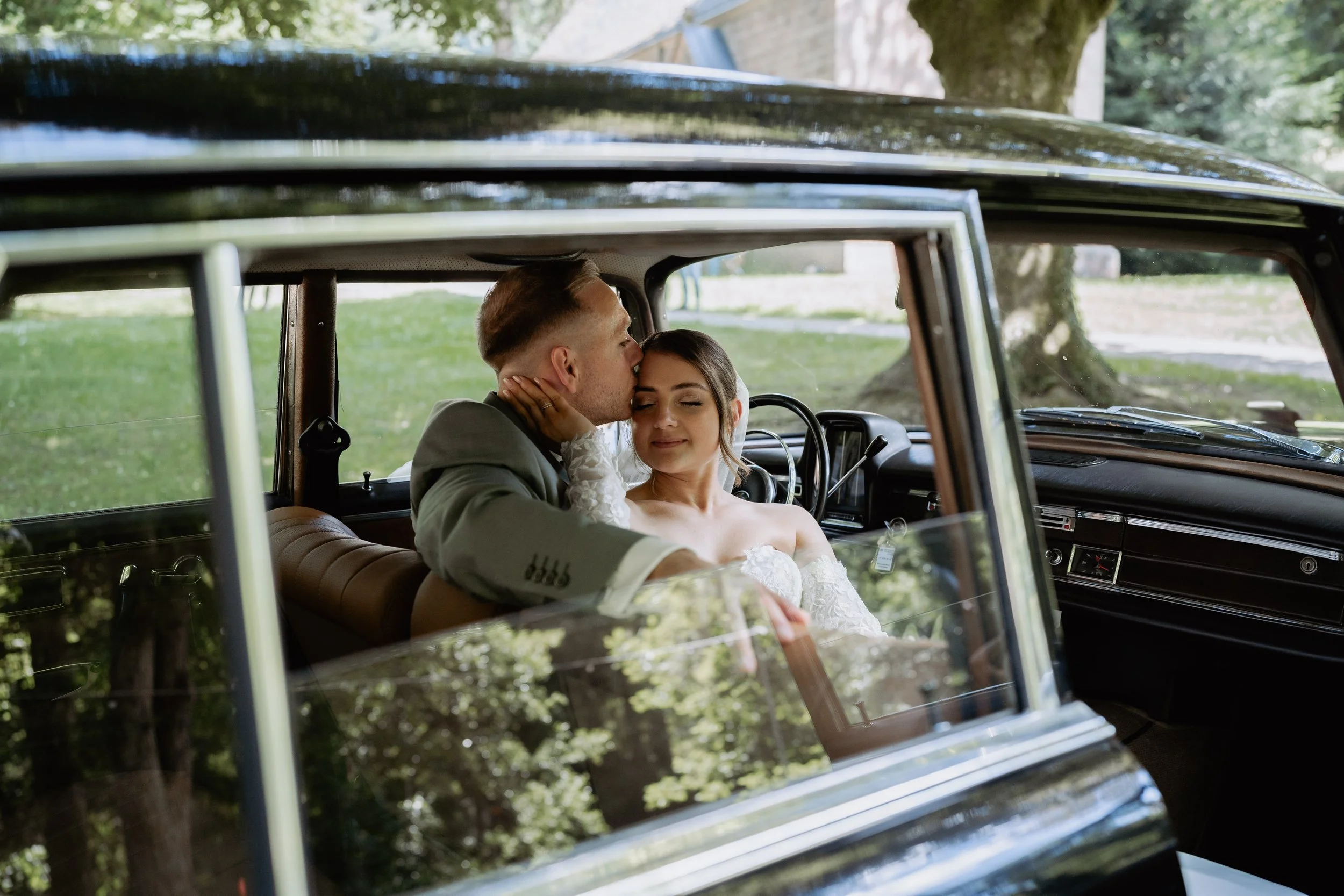 A bride and groom sitting inside a vintage car with the groom kissing the bride on the forehead during daytime.