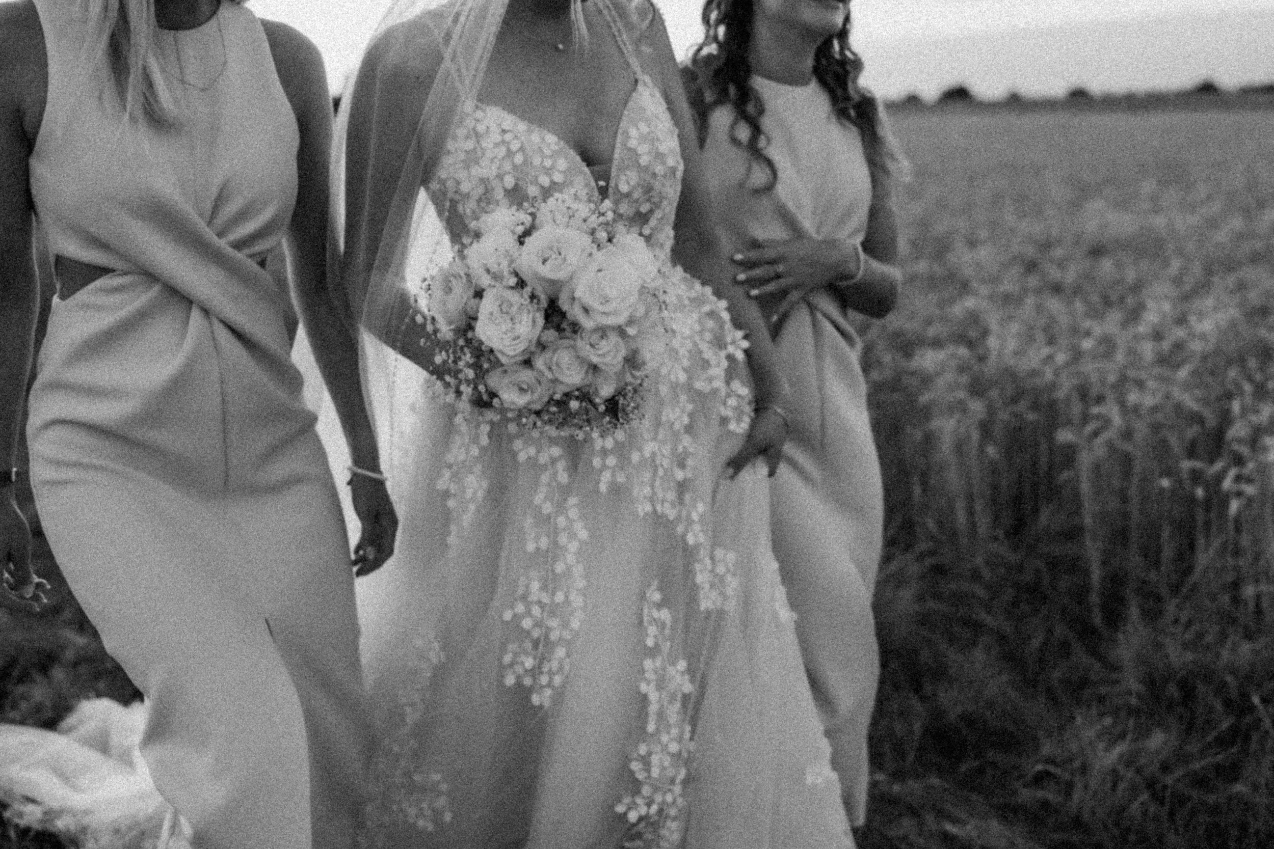 Three women standing outdoors, one in a wedding dress holding a bouquet, and two in dresses, with a grassy field and trees in the background.
