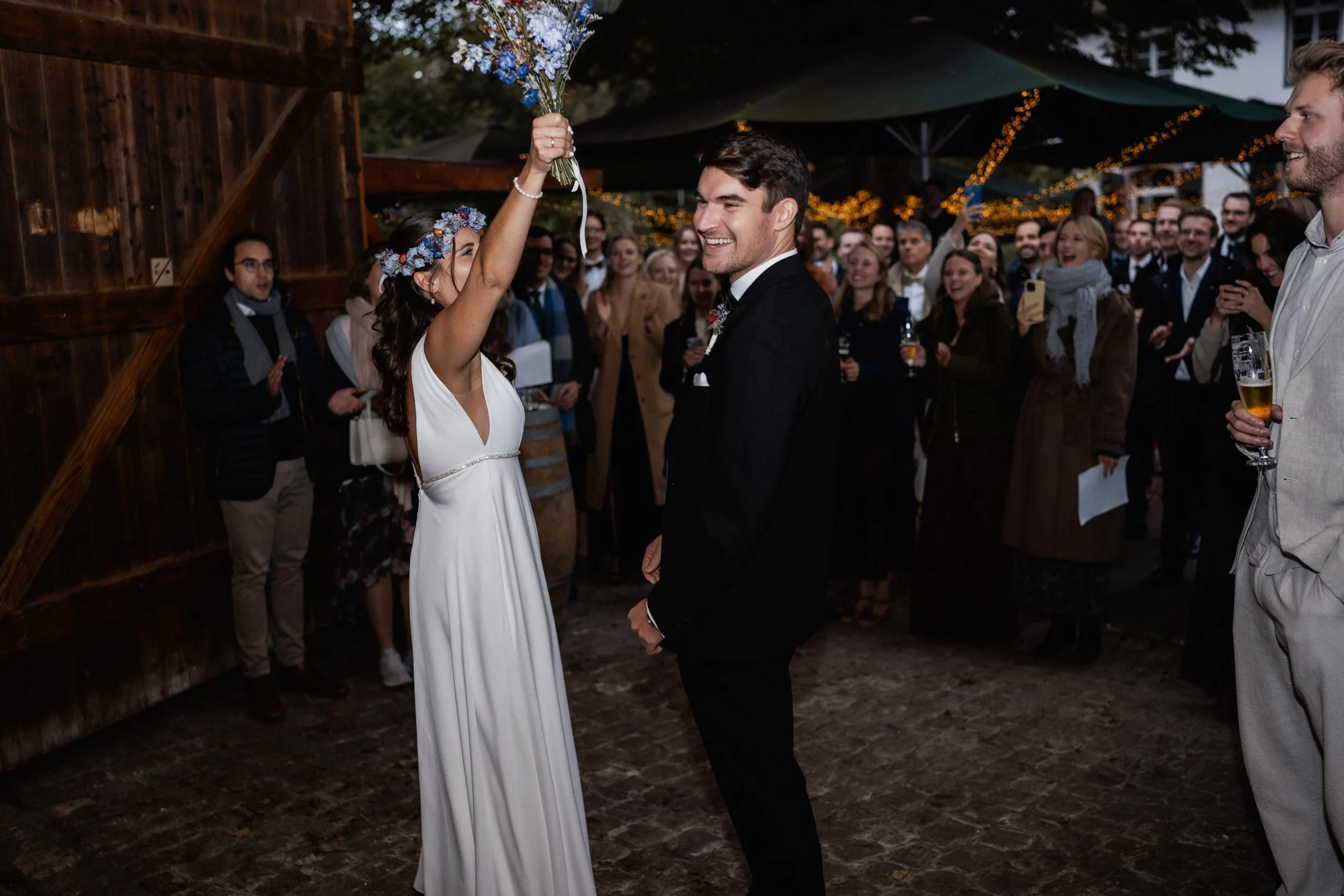 A bride and groom share a tender moment at their wedding reception, with the bride holding a bouquet above her head and both smiling, surrounded by a gathering of guests under string lights and outdoor canopies.