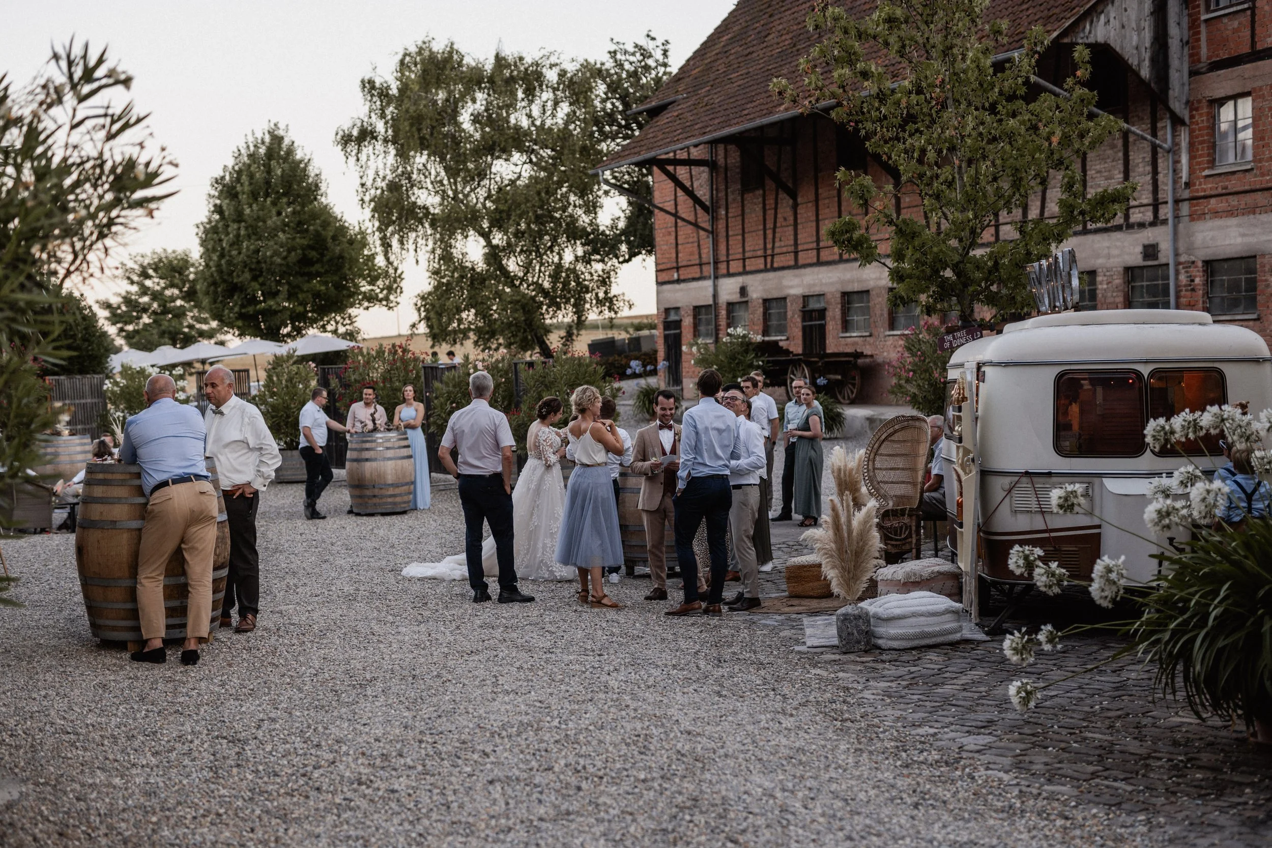 People gather outdoors in front of a rustic brick building during a wedding reception, with some standing and talking, while the bride and groom share a moment in the center, surrounded by greenery and decorative elements.