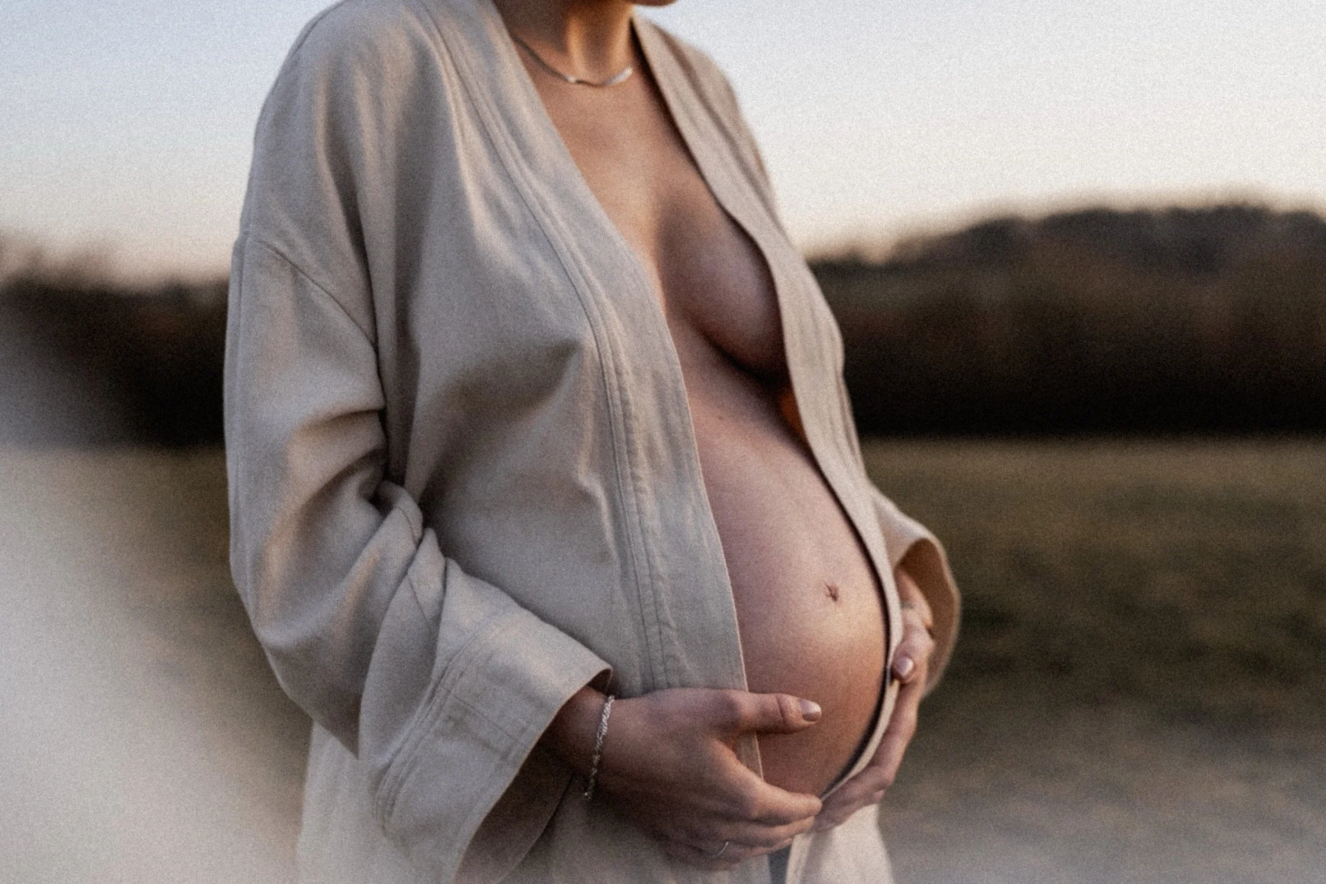 A person with a bare abdomen gently cradles their belly outdoors at sunset, wearing an open beige jacket and a silver necklace.